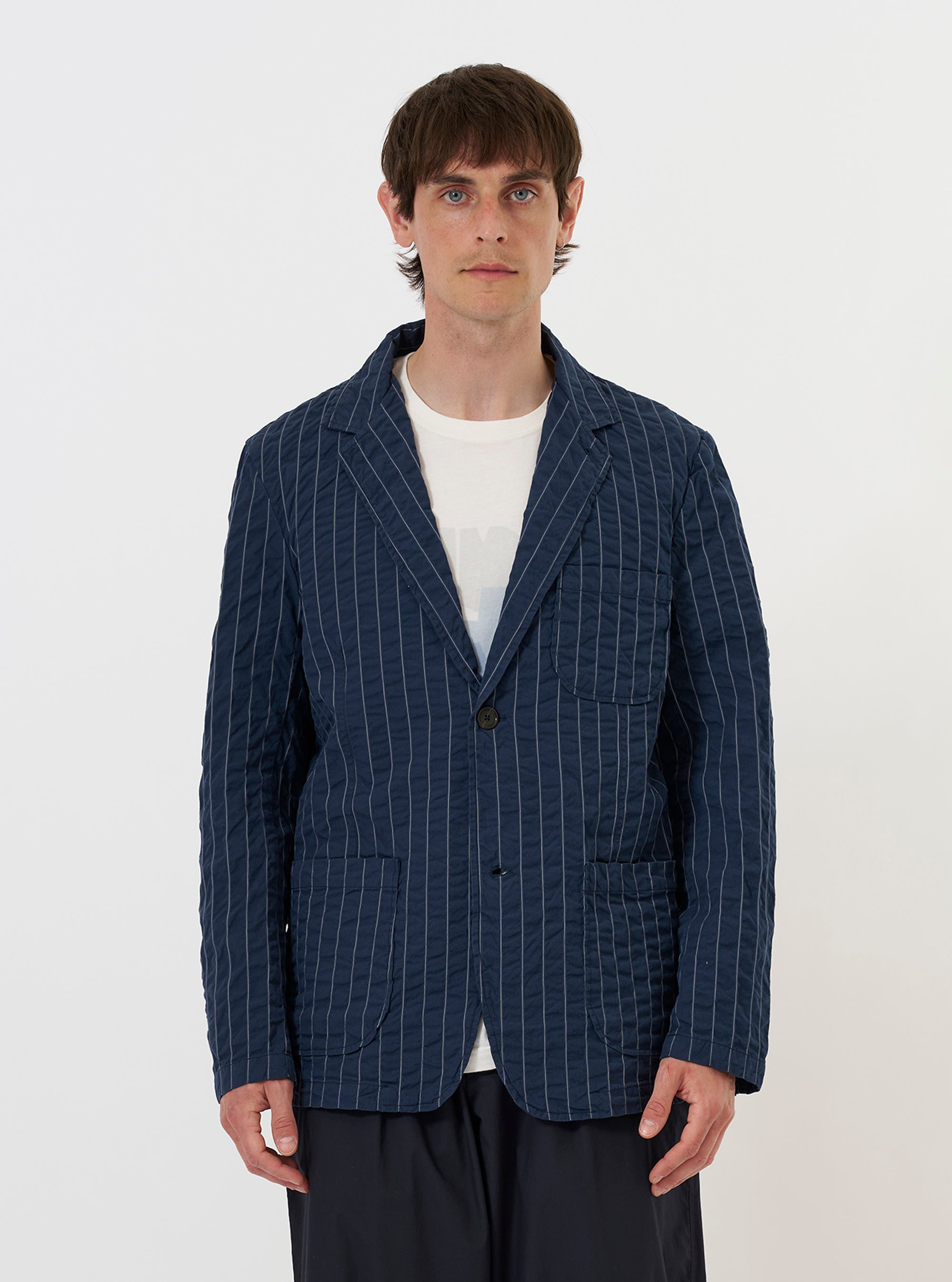 A model is wearing the Navy Chester Stripe Two Button Jacket against a plain white background. The jacket features a relaxed fit with a navy and white seersucker stripe pattern, a long and wide lapel, two lower patch pockets, and a single chest patch pocket. The model is shown from the waist up, and the jacket is worn over a light-colored shirt. No mannequin is present in the image.
