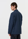 A model stands against a plain white background wearing the Navy Chester Stripe Two Button Jacket. The image is taken from a side-rear perspective, highlighting the jacket's relaxed fit and the vertical white stripes on the navy cotton fabric. The model is shown from the chest down, wearing the jacket over a matching garment.