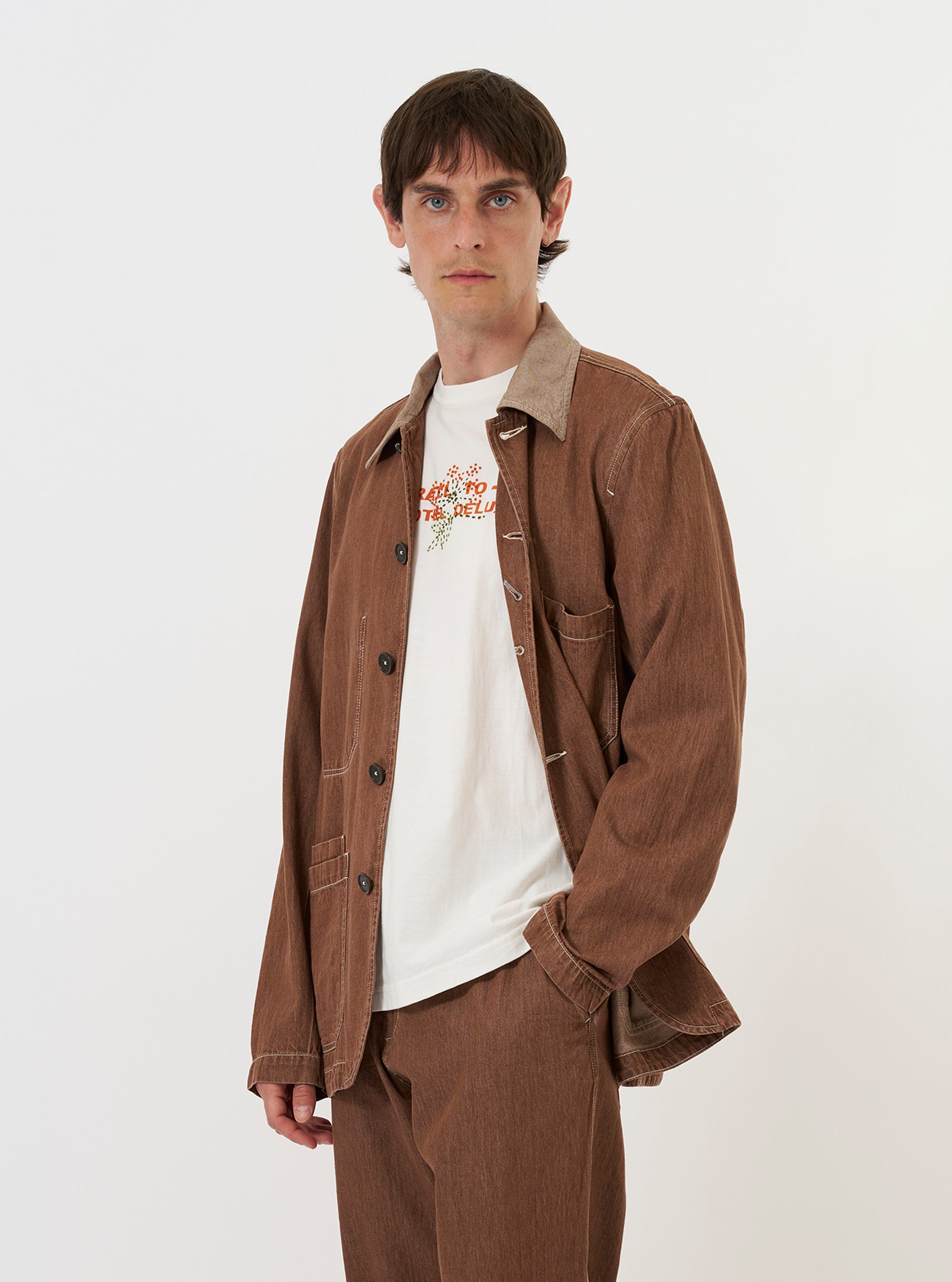 A model is wearing the Brown Meco Denim OG Bakers Jacket against a plain white background. The brown denim chore jacket features a loose fit, a classic collar, and multiple front patch pockets with contrast stitching. The model is shown from the waist up, wearing the jacket over a light-colored shirt.