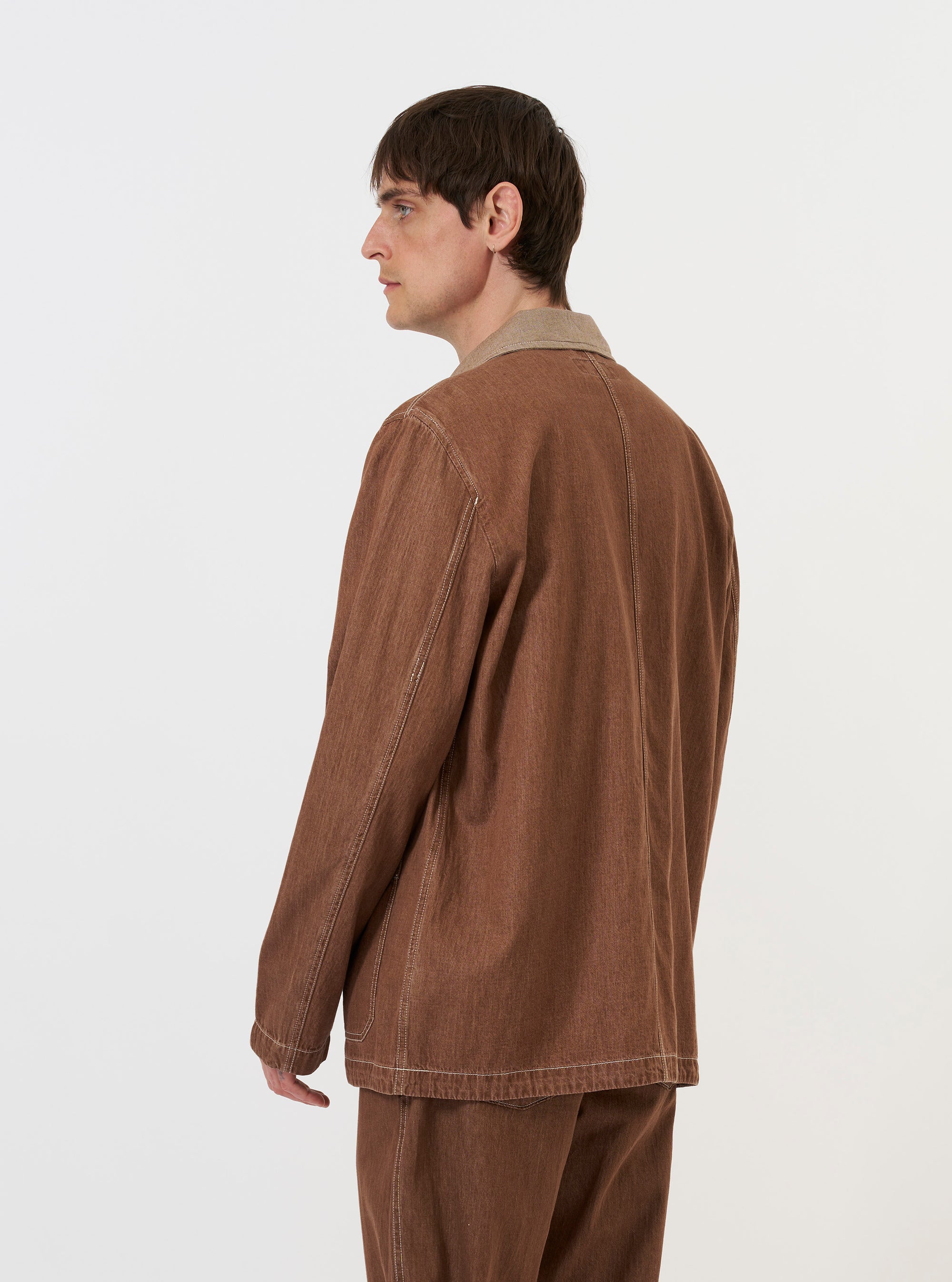 A model is wearing the Brown Meco Denim OG Bakers Jacket against a plain white background. The image, captured from a side-rear perspective, highlights the jacket's loose fit, the central back seam, and the two-piece sleeve construction. The model is standing with their hands by their sides, showcasing the length of the garment and the texture of the brown recycled cotton denim. No mannequin is present in the image.