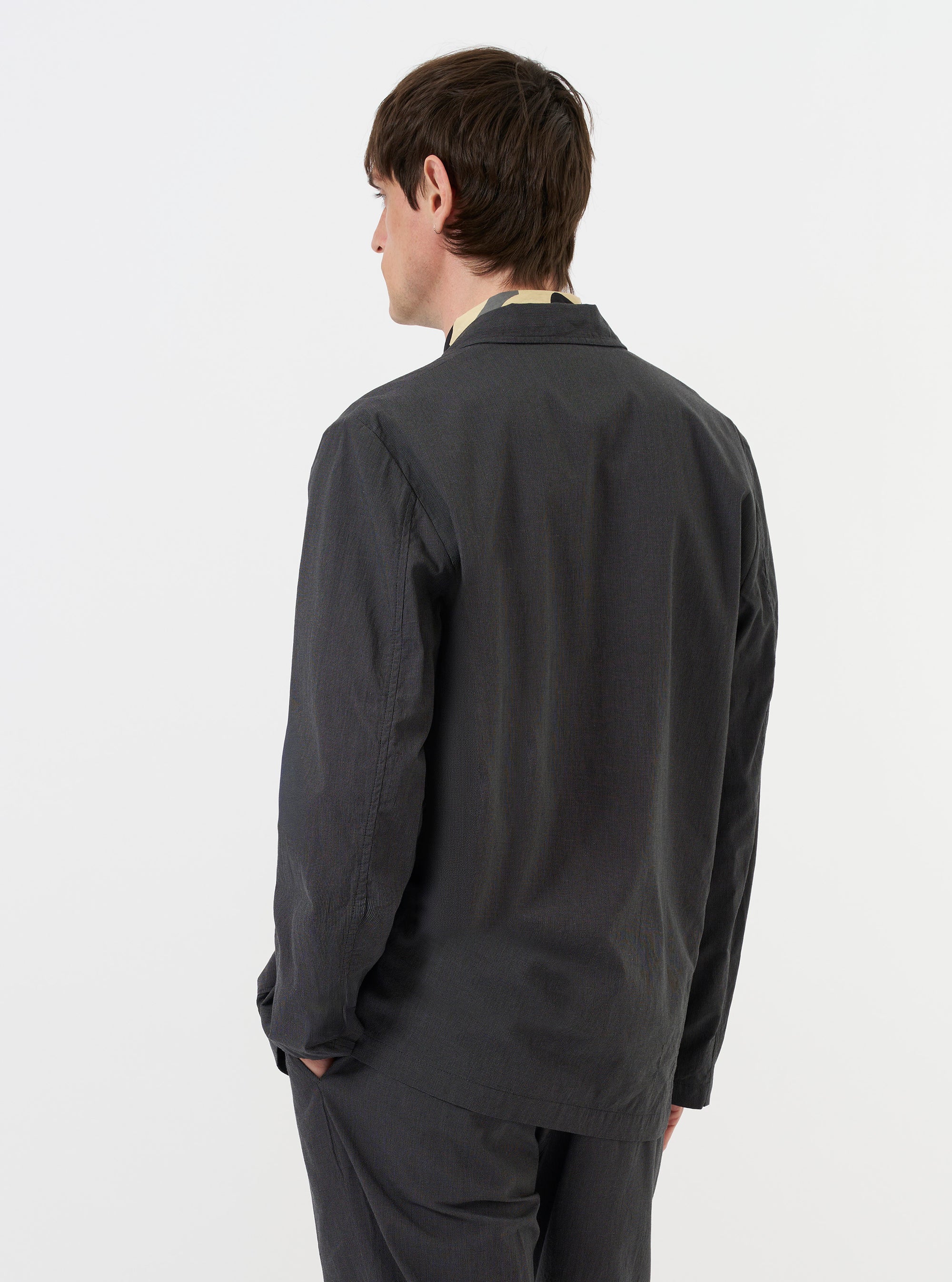 A model is seen from the back wearing the Charcoal Seattle Cotton Two Button Jacket against a plain white background. The image highlights the jacket's relaxed fit, structured shoulders, and central vent at the hem. The charcoal grey cotton fabric features a subtle basket-weave texture.