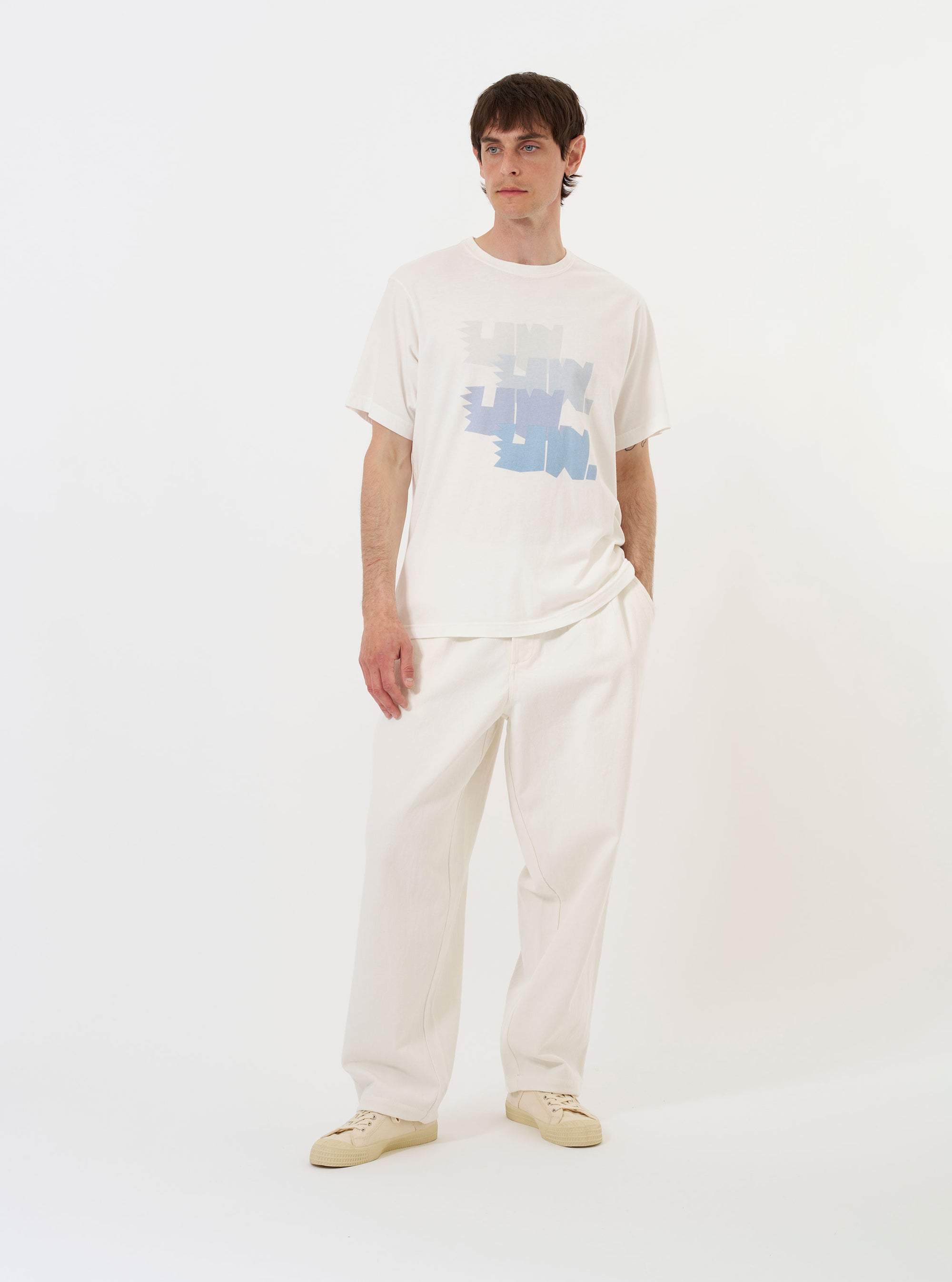 The Ecru Single Jersey Multi UW Print Tee is shown laid flat against a solid white background. The short-sleeved t-shirt features a classic crew neck and a prominent multi-colored graphic print centered on the chest, consisting of four graduated rows of the letters "UW" in shades of blue, orange, and navy.