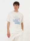 A model stands against a plain white background wearing the Ecru Single Jersey Multi UW Print Multi UW Print Tee. The short-sleeved t-shirt features a repetitive black graphic print across the ecru-colored fabric and a classic crew neck. The model is shown from the waist up, highlighting the relaxed fit and the prominent all-over pattern.