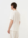 A model is wearing the Ecru Single Jersey Multi UW Print Multi UW Print Tee against a plain white background. The image is taken from a side-rear perspective, showcasing the relaxed fit, drop shoulder, and the repeating navy blue logo print across the ecru-colored fabric.