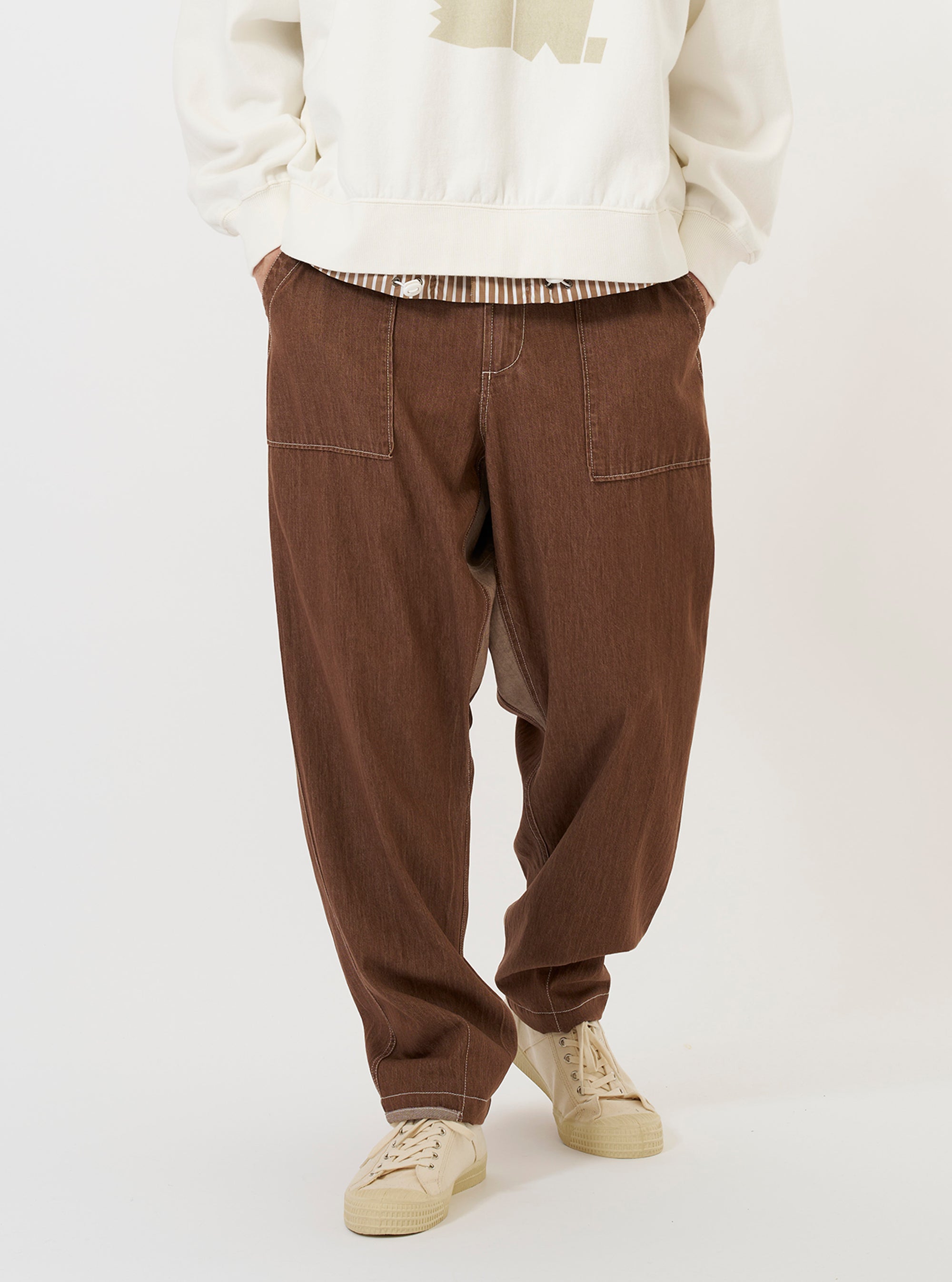 A model wears a pair of Brown Meco Denim Patched Fatigue Pants while standing against a plain white background. The trousers feature a straight-leg fit, large rectangular patch pockets on the front of the thighs, and a visible button fly. The model is also wearing a light-colored shirt and navy blue shoes.