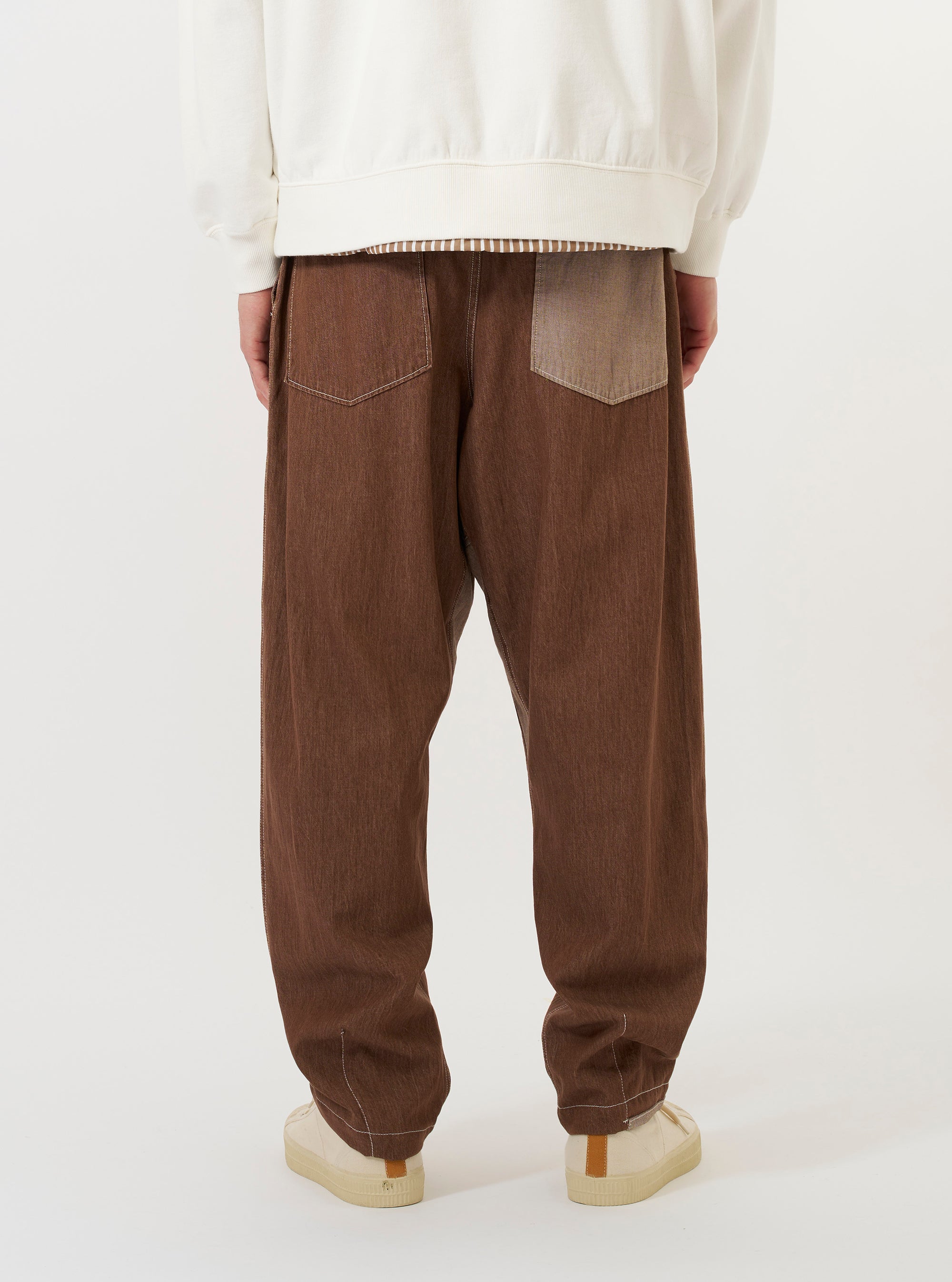 A model is wearing a pair of Brown Meco Denim Patched Fatigue Pants against a plain white background. The image provides a close-up view from the waist down, showcasing the straight-leg fit, large fatigue-style front patch pockets, and the texture of the brown denim. The model is standing and is also wearing navy blue footwear.