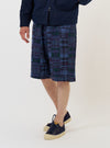 A model stands against a plain white background wearing the Navy Madras Patchwork Vacation Shorts. The shorts feature a vibrant blue and navy plaid patchwork pattern and a relaxed fit. The model is also wearing navy shoes, and the image captures the garment from the waist down, highlighting the texture and drape of the fabric.