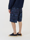 A model wears the Navy Madras Patchwork Vacation Short, featuring a blue and white plaid pattern, against a plain white background. The image highlights the side view of the shorts, showing the relaxed fit and mid-thigh length, paired with navy blue shoes. No mannequin is present.