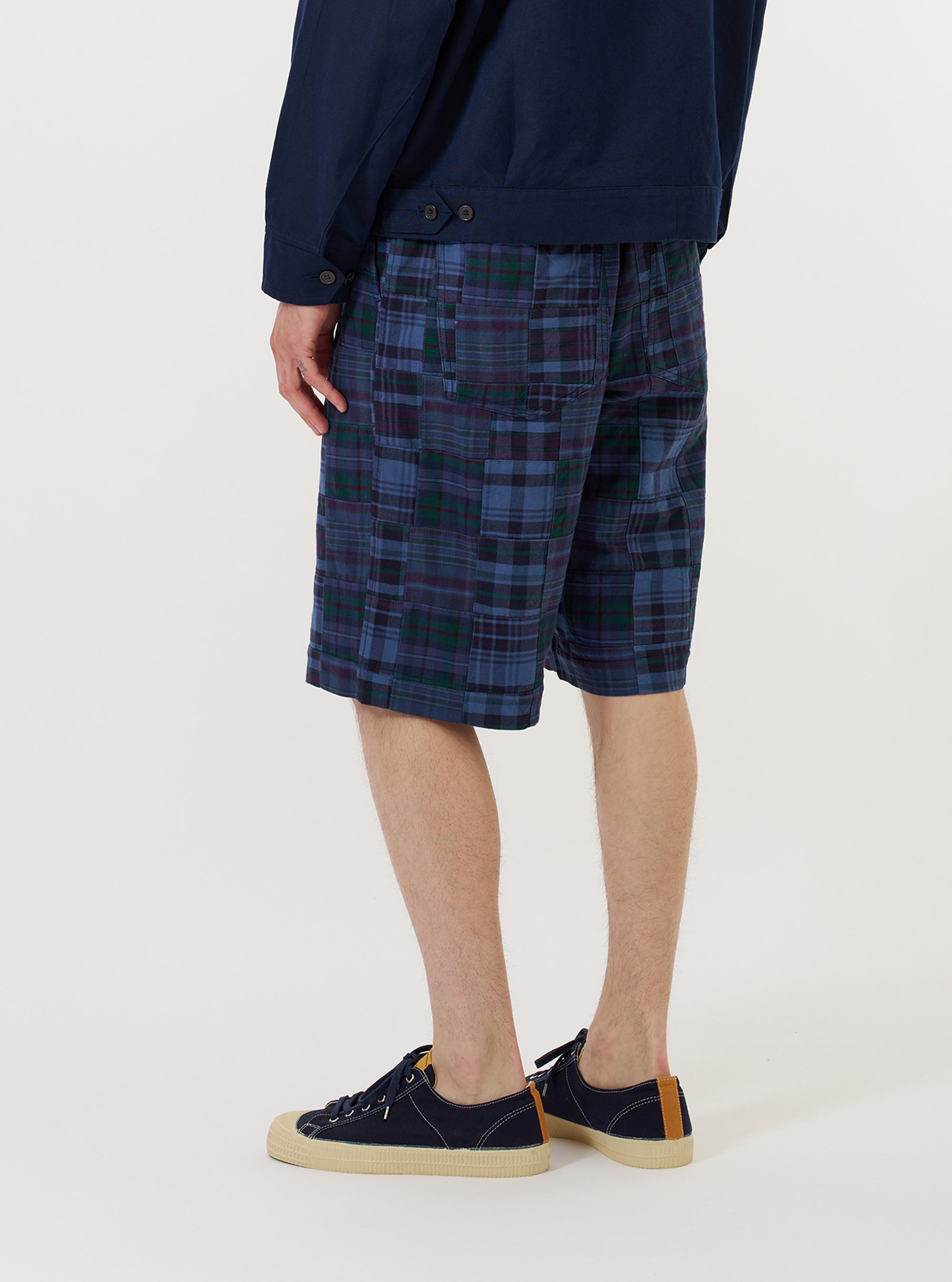 A model wears the Navy Madras Patchwork Vacation Short, featuring a blue and white plaid pattern, against a plain white background. The image highlights the side view of the shorts, showing the relaxed fit and mid-thigh length, paired with navy blue shoes. No mannequin is present.