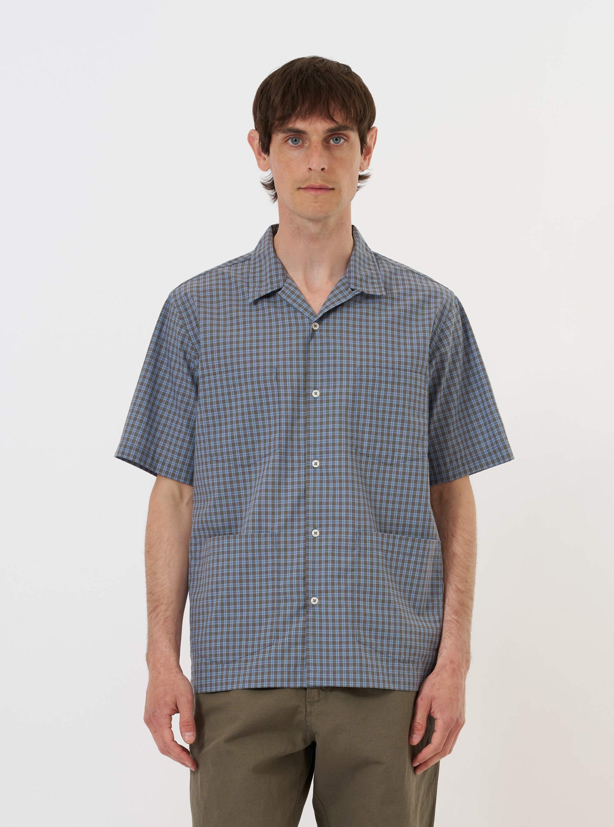 A pale blue check plaid summer overshirt is displayed flat against a plain white background. The garment features a standard pointed collar, a full button-down front, and two prominent utility chest pockets with buttoned flaps. The fabric has a light blue and white plaid pattern.