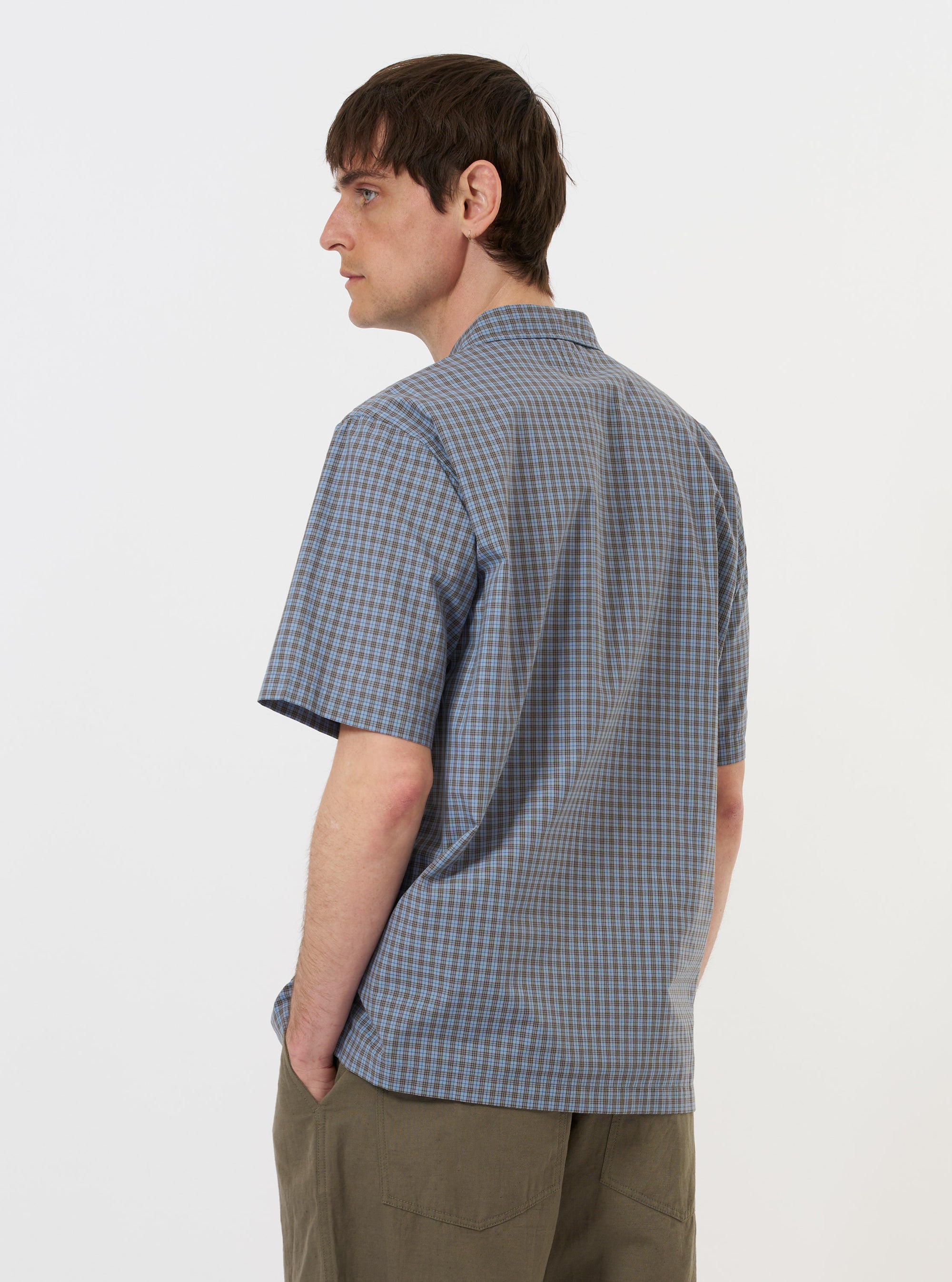 A model is seen from the back wearing the Pale Blue Check Plaid Summer Overshirt against a plain white background. The overshirt features a light blue and white plaid pattern, a straight hem, and a simple collar. The model is also wearing dark trousers.