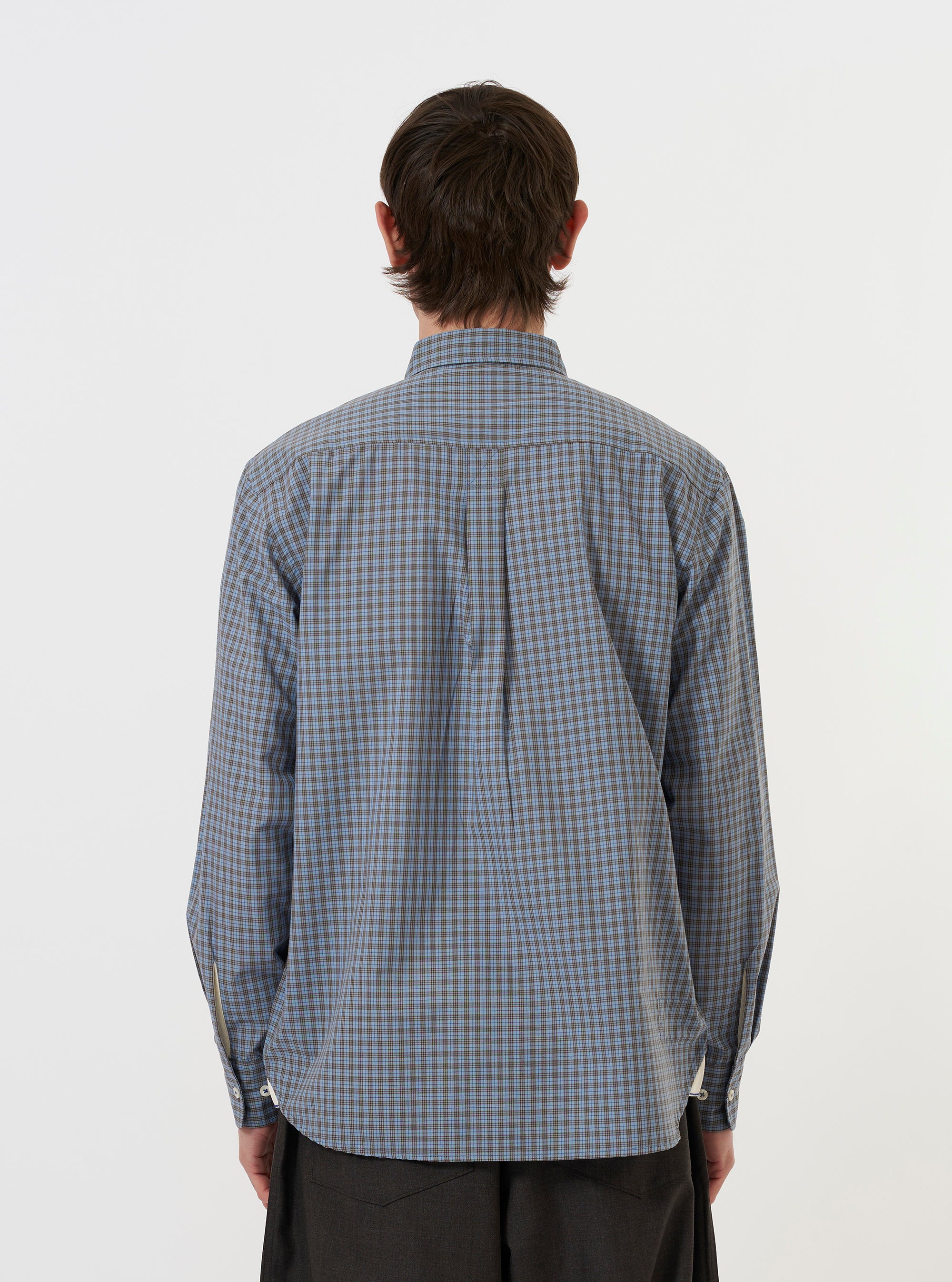A model is wearing the Pale Blue Check Plaid Lazy Day Shirt, standing against a plain white background. The image shows the shirt from the back, highlighting the yoke, the hang loop, and the subtle texture of the pale blue plaid pattern. The model has their hands in their pockets, and the shirt is paired with dark trousers.