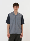 A model wears the Mixed Check Plaid Camper Shirt against a plain white background. The shirt features a multi-colored patchwork design with various plaid patterns, short sleeves, and a relaxed camp collar. It has two prominent chest pockets and is worn open over a light-colored t-shirt.