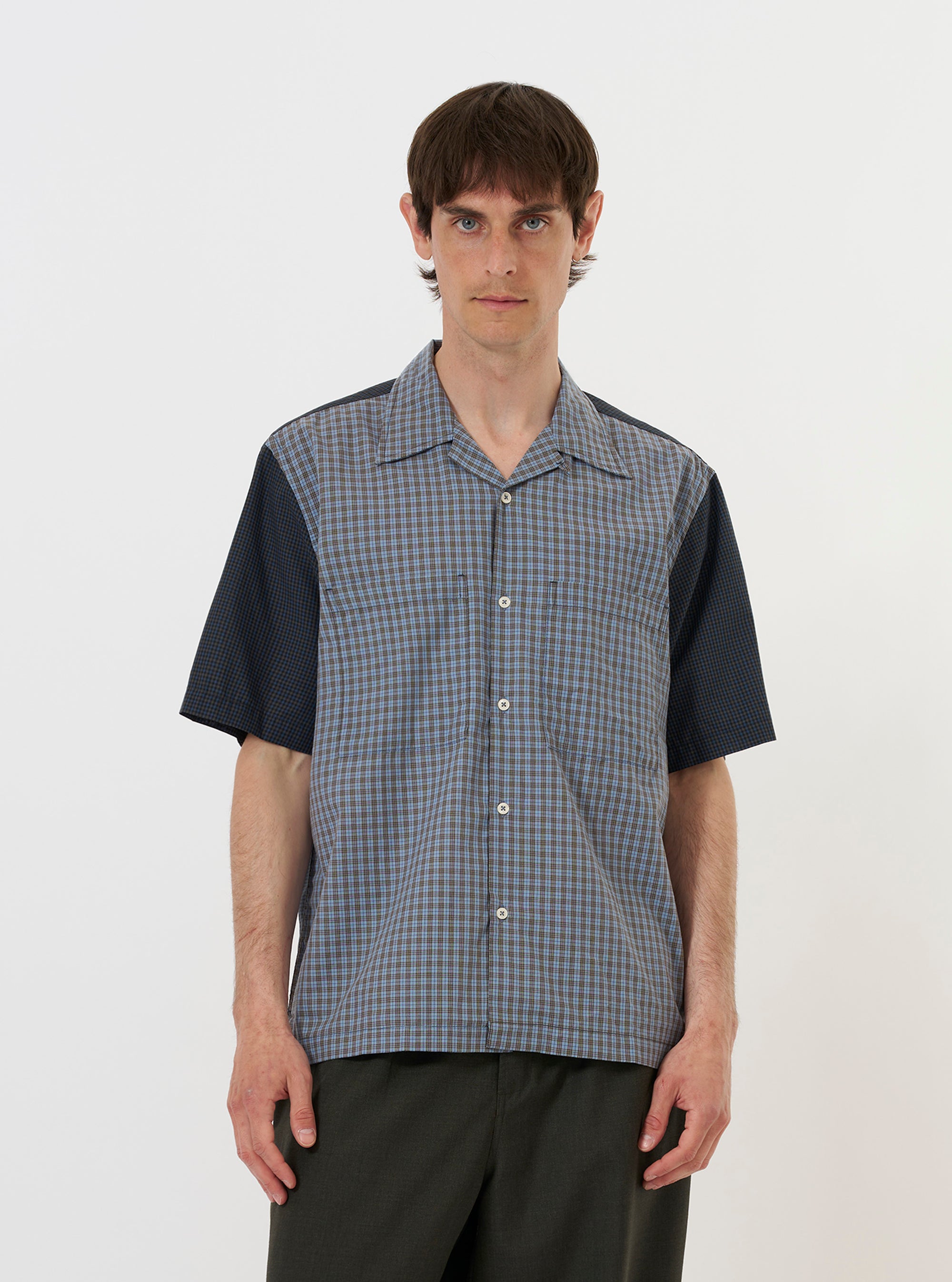 A model wears the Mixed Check Plaid Camper Shirt against a plain white background. The shirt features a multi-colored patchwork design with various plaid patterns, short sleeves, and a relaxed camp collar. It has two prominent chest pockets and is worn open over a light-colored t-shirt.