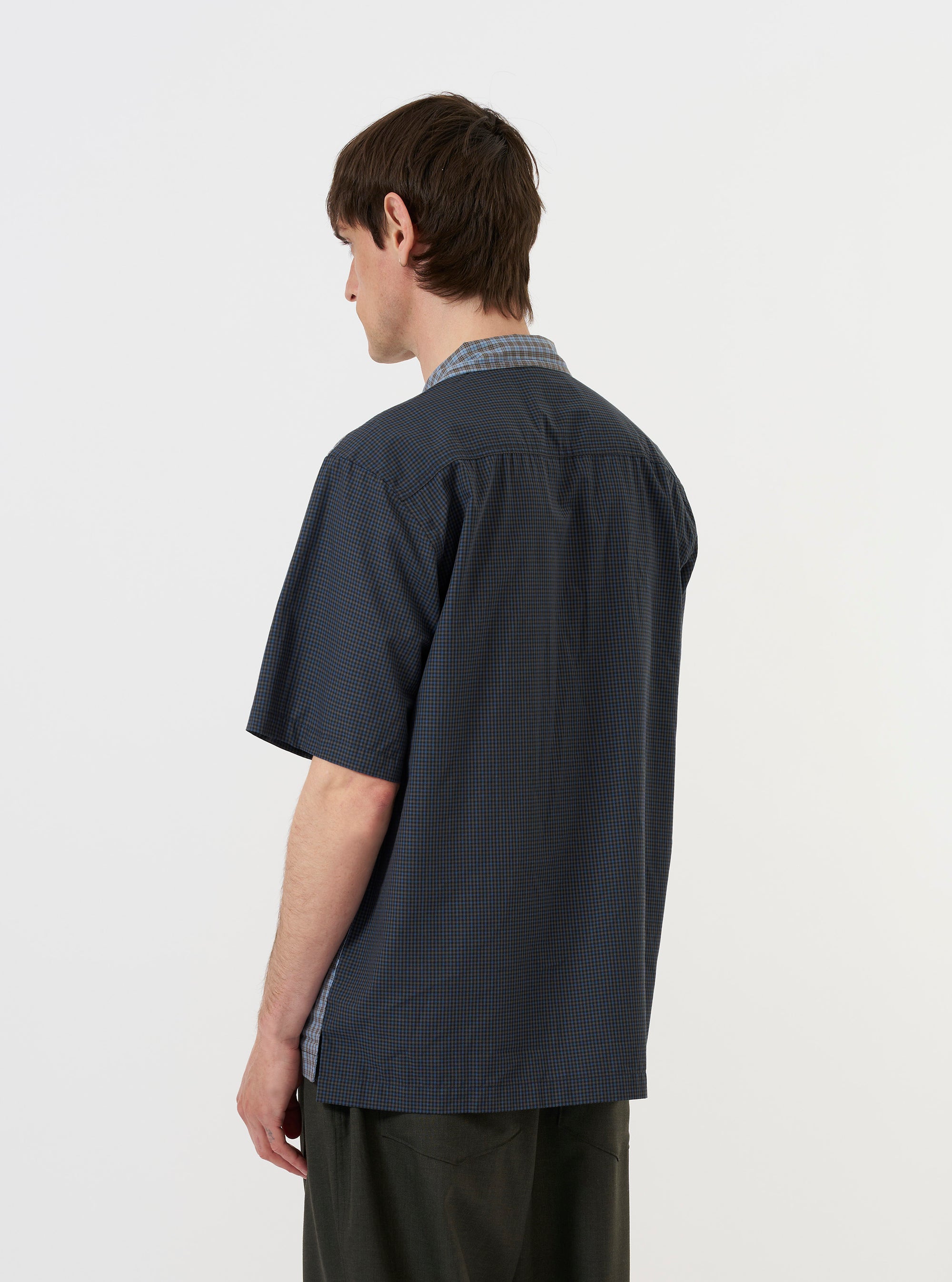 A model wears a Mixed Check Plaid Camper Shirt, featuring a multi-colored patchwork of blue, cream, and orange plaid patterns. The short-sleeved shirt is styled with an open camp collar and a button-down front, and is worn over a dark navy undershirt. The model is photographed against a plain, light grey background.