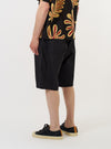 A model is wearing the Black Broadcloth Vacation Short against a plain white background. The image provides a side-rear view of the black shorts, highlighting the relaxed fit, mid-thigh length, and a single patch pocket on the back. The model is standing and paired the shorts with dark footwear.