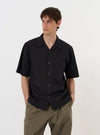 The Black Broadcloth Camp Shirt is displayed flat against a plain white background. The garment features a short-sleeved, button-down design with a distinctive one-piece camp collar and a large patch pocket on the left chest. The shirt is made from a smooth black broadcloth fabric and has a straight hem.