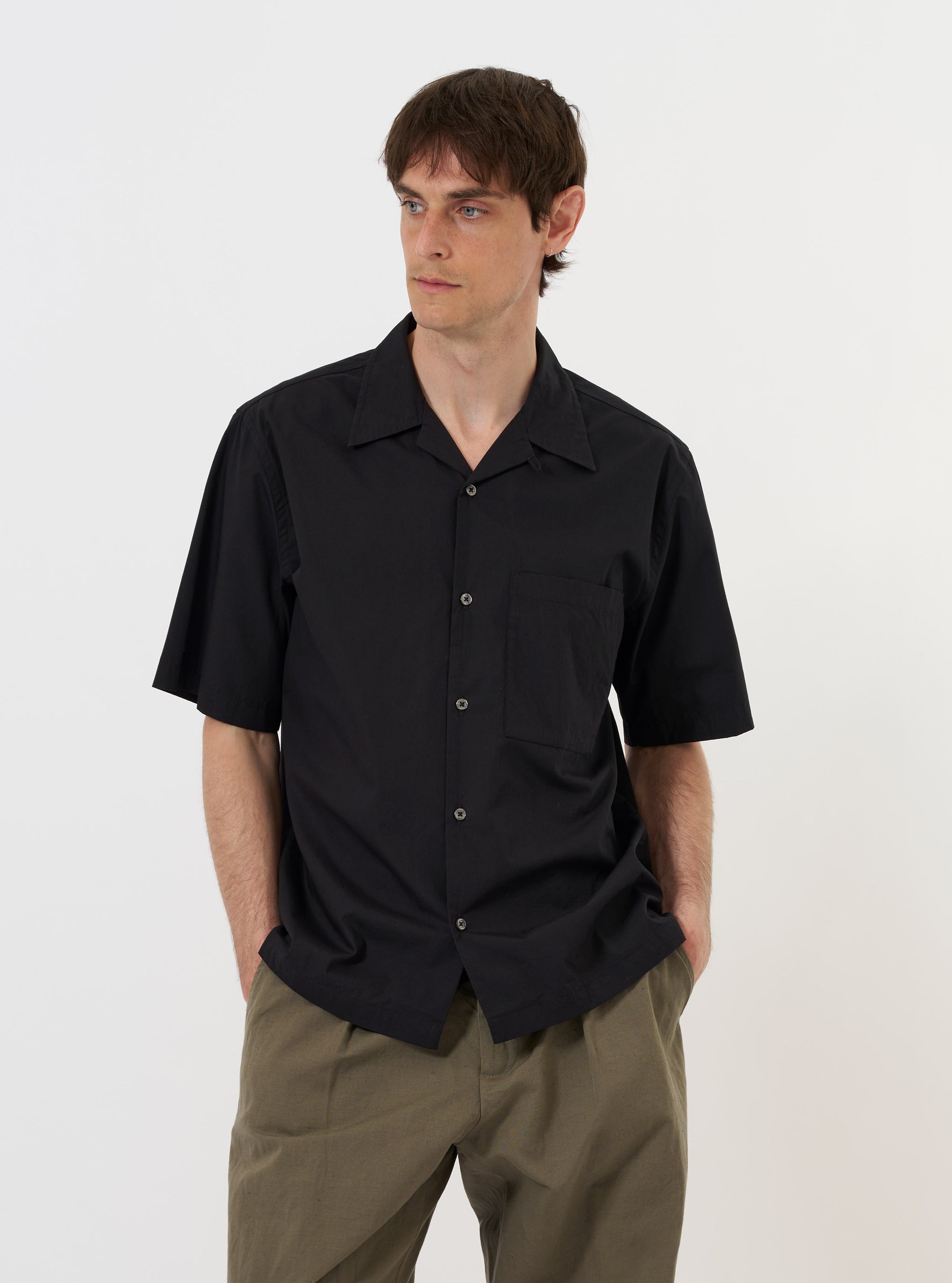 The Black Broadcloth Camp Shirt is displayed flat against a plain white background. The garment features a short-sleeved, button-down design with a distinctive one-piece camp collar and a large patch pocket on the left chest. The shirt is made from a smooth black broadcloth fabric and has a straight hem.