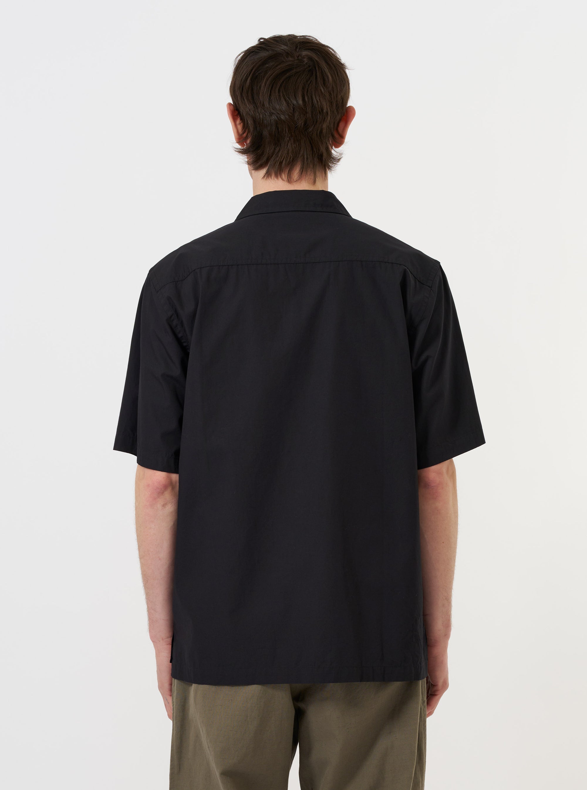 The Black Broadcloth Camp Shirt is shown from the back, displayed flat against a plain white background. The image highlights the horizontal back yoke with two small pleats for added volume and the straight hem with side splits.