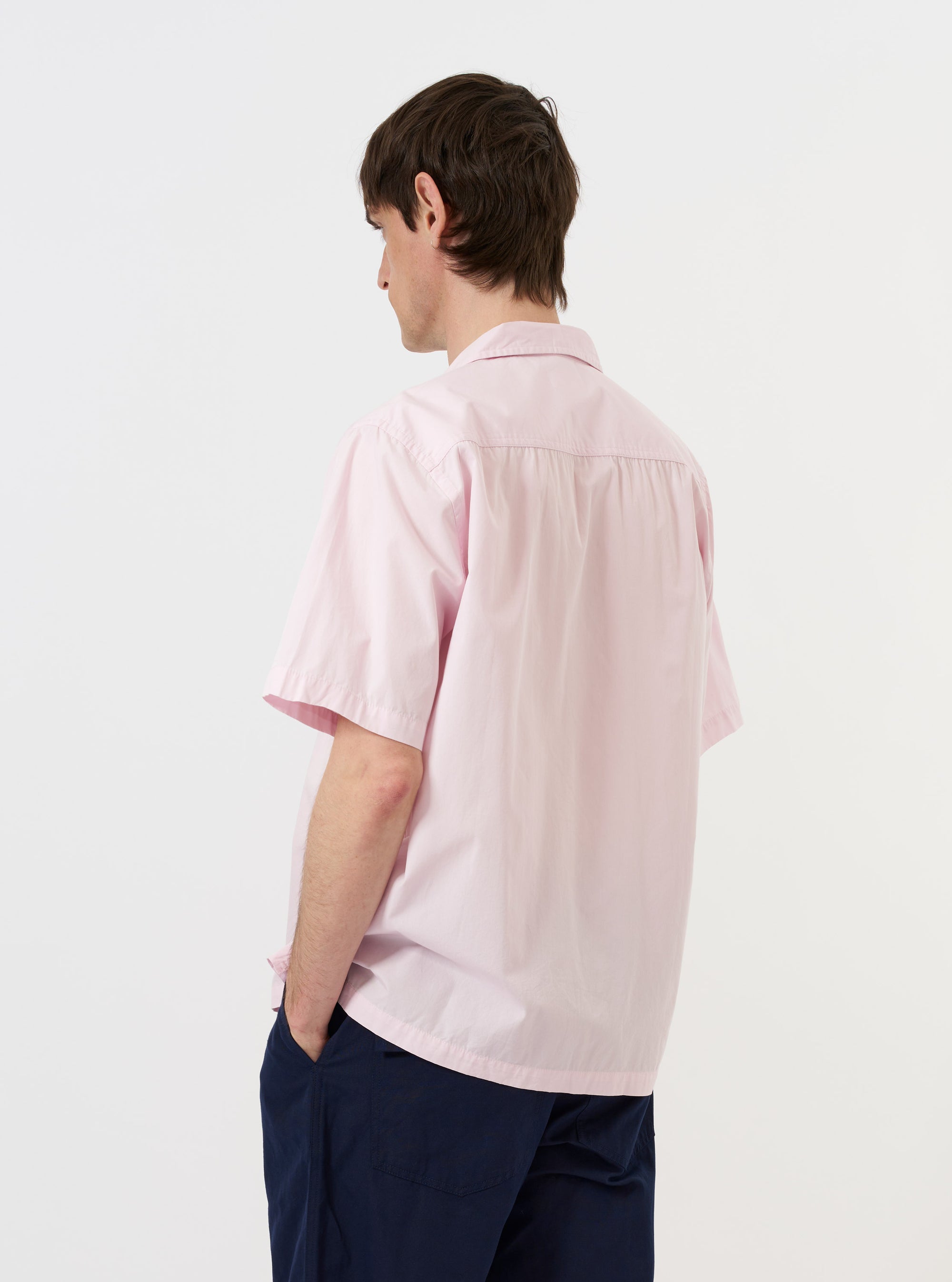 A model is seen from the back wearing the Pale Pink Broadcloth Camp Shirt against a plain white background. The short-sleeved shirt features a relaxed fit, a straight hem, and a simple back yoke. The model is also wearing dark trousers.
