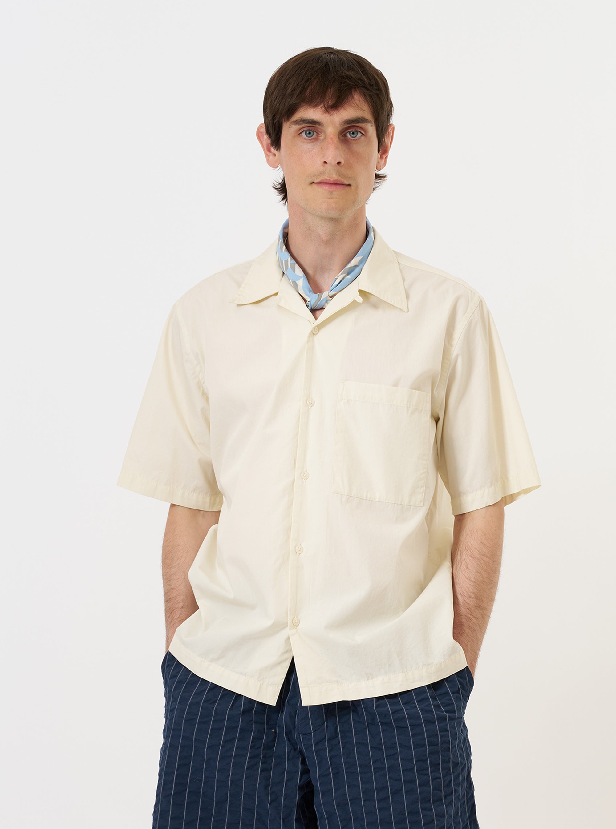 A model is wearing the Ecru Broadcloth Camp Shirt while standing against a plain white background. The image highlights the front of the short-sleeved shirt, featuring a relaxed wide fit, a one-piece camp collar, and a single large square chest pocket. The model is wearing the shirt buttoned and untucked.