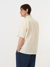 A model is wearing the Ecru Broadcloth Camp Shirt against a plain white background. The image shows the shirt from the back, highlighting the clean, smooth texture of the ecru cotton broadcloth, a straight hem, and a simple back yoke. The model is standing with their arms at their sides, and the shirt is paired with dark trousers. No mannequin is present in the image.