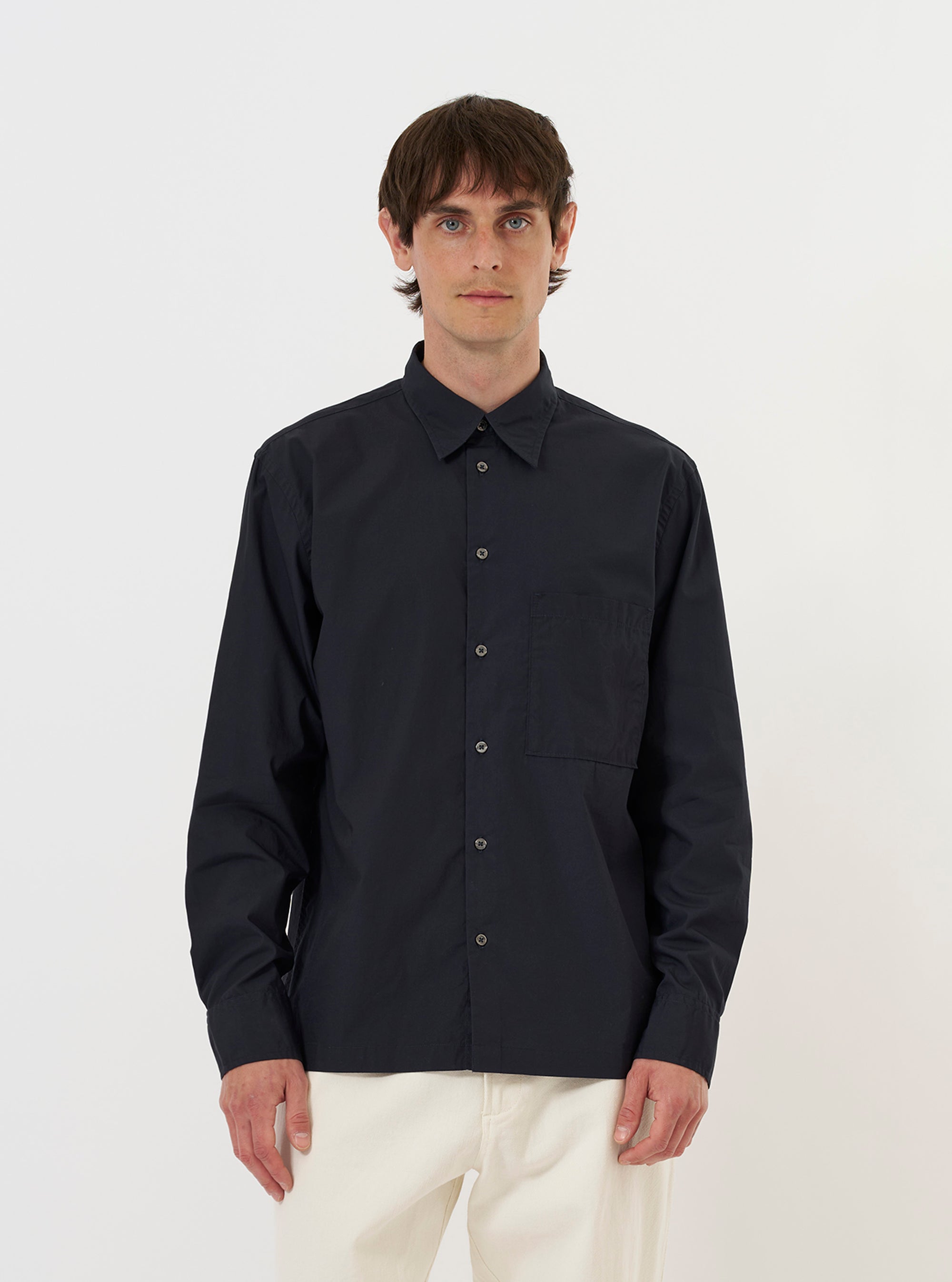 A model is wearing the Navy Broadcloth Lucky Shirt against a plain white background. The navy blue shirt features a relaxed fit, a classic pointed collar, and a full button-down front. The model is shown from the waist up, wearing the garment over a light-colored t-shirt, highlighting the smooth texture of the broadcloth fabric.