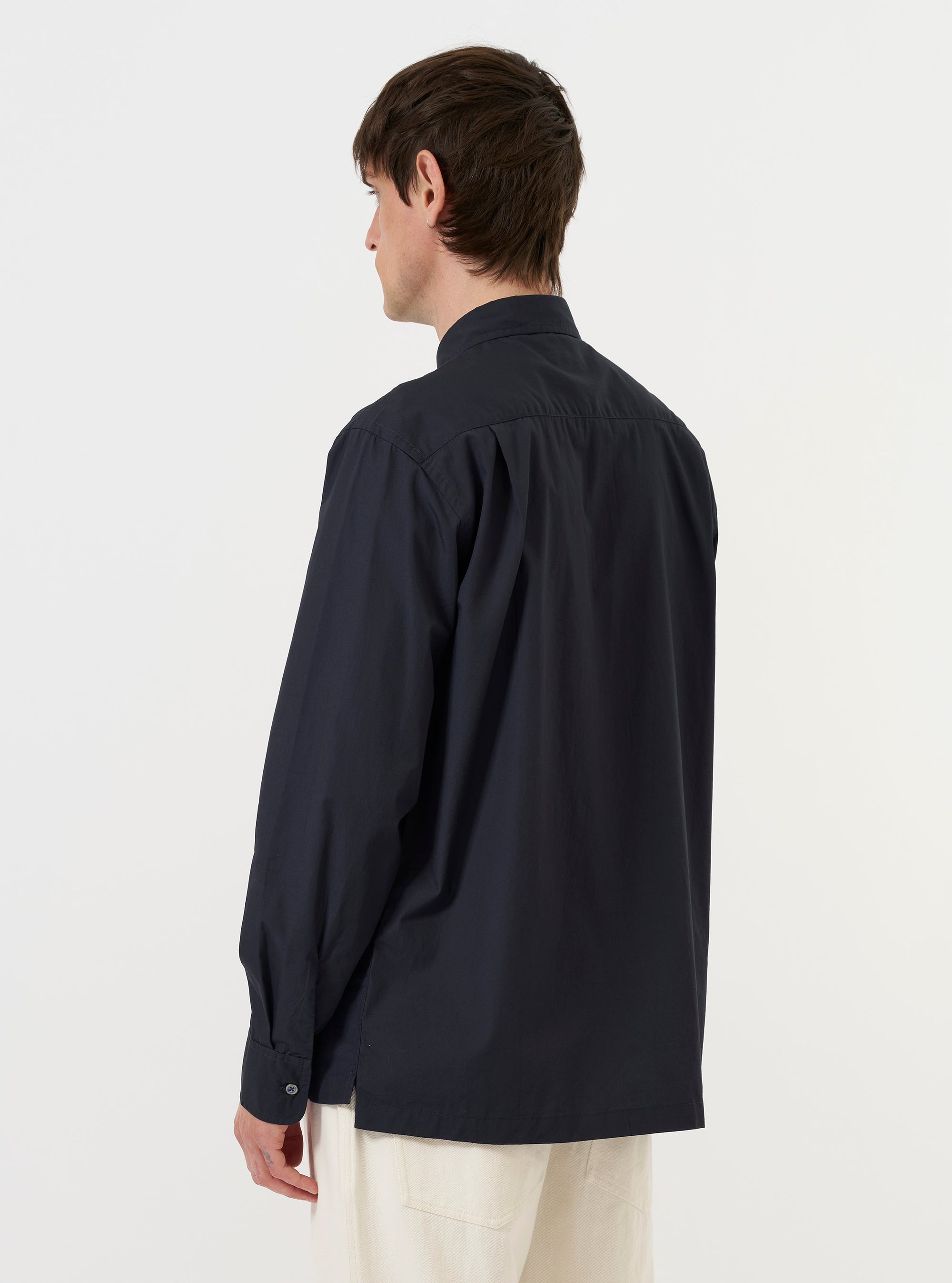 A model is wearing the Navy Broadcloth Lucky Shirt against a plain white background. The image captures the garment from the back, showcasing the straight yoke and the smooth texture of the navy blue broadcloth. The shirt is worn untucked over dark trousers.