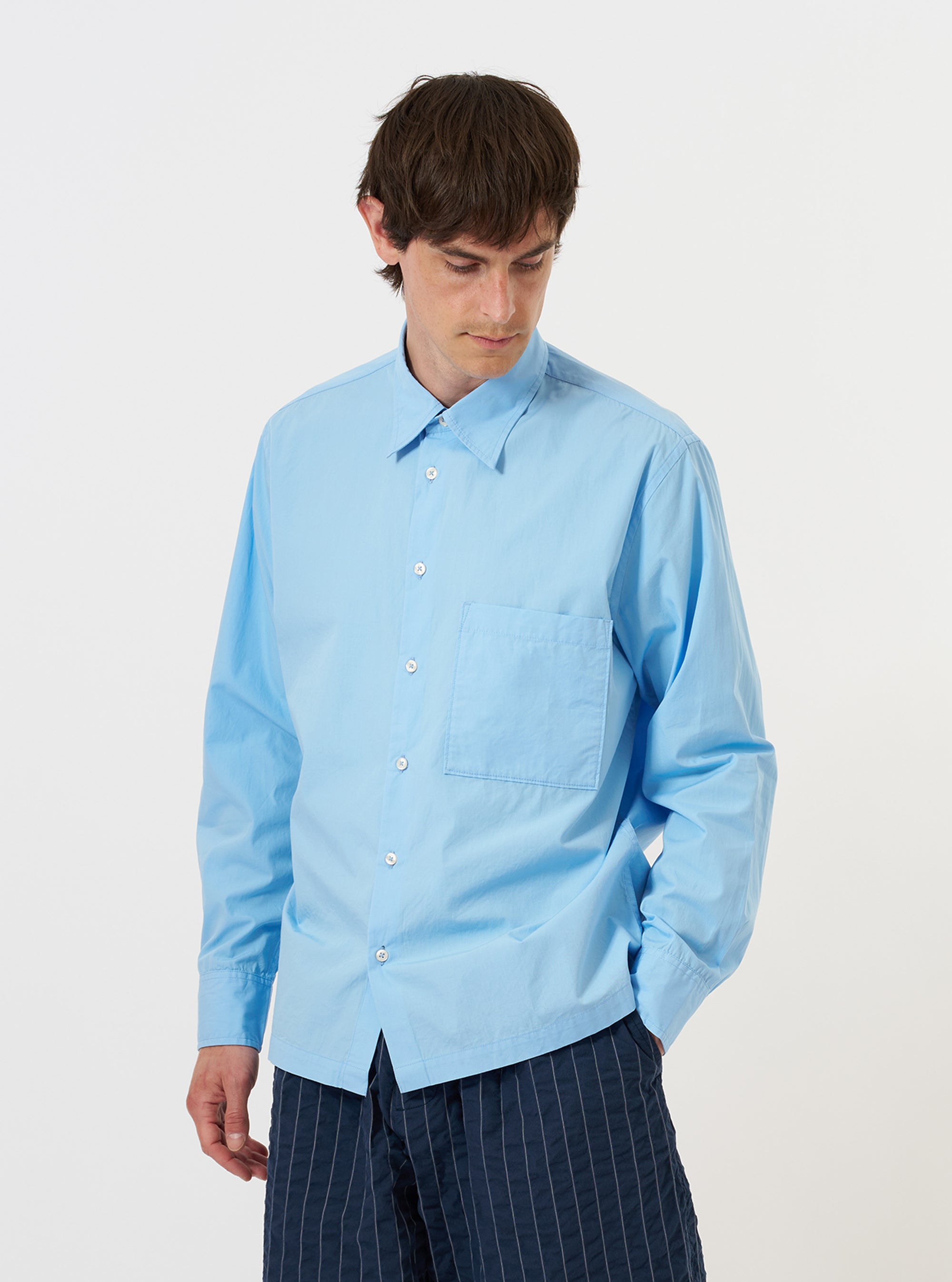 A model is wearing the Pale Blue Broadcloth Lucky Shirt against a plain white background. The shirt features a pale blue color, a classic collar, and a full button-down front. The model is shown from the torso up, wearing the garment over a white t-shirt, highlighting the relaxed fit and the texture of the broadcloth fabric.