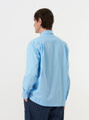 A model is seen from the back wearing the Pale Blue Broadcloth Lucky Shirt against a plain white background. The image highlights the shirt's relaxed fit, a horizontal back yoke, and a central box pleat. The model has their hands in their pockets and is wearing dark trousers. No mannequin is present in the image.