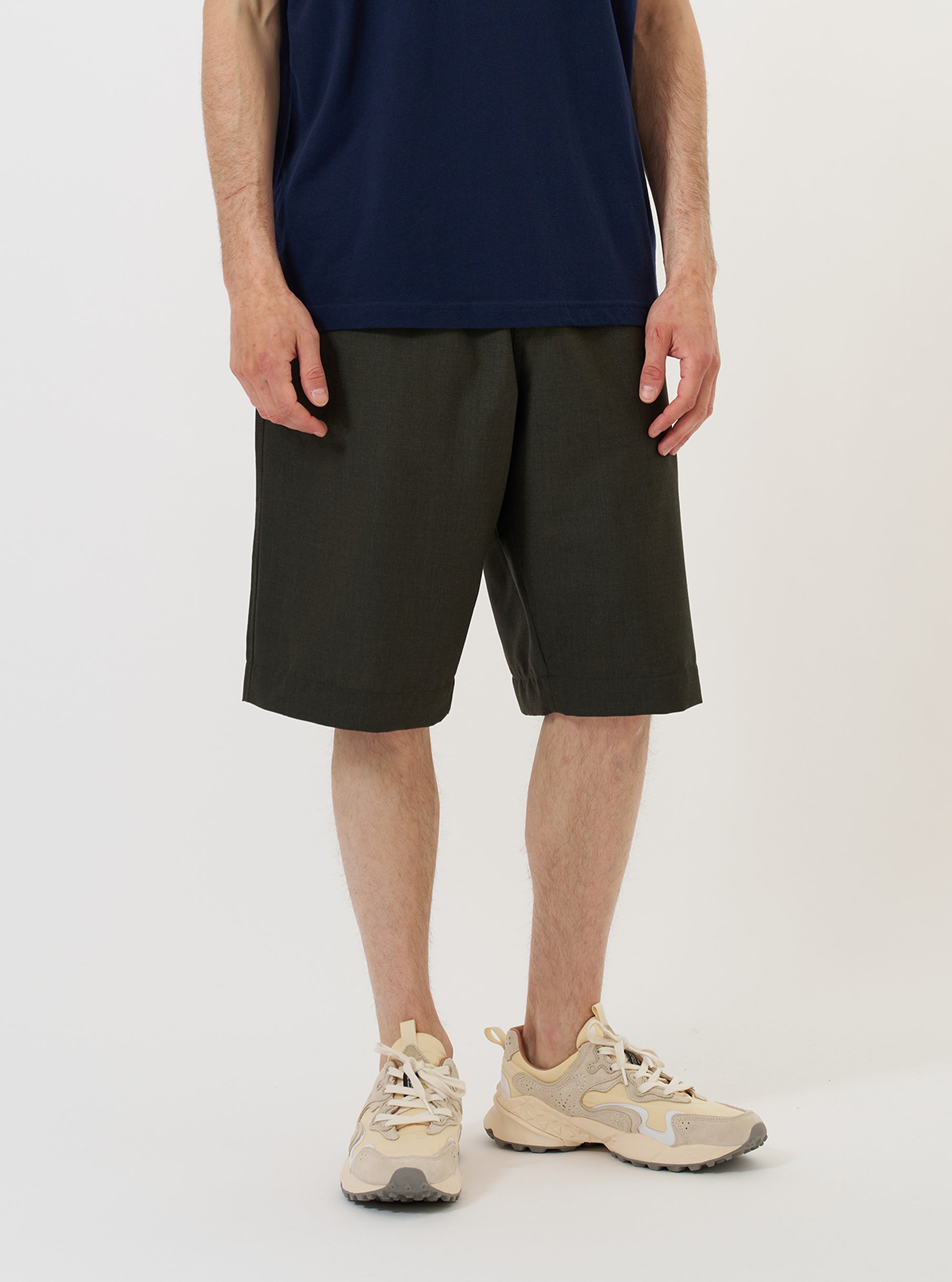 A model stands against a plain white background wearing the Olive Marl Tropical Suiting Vacation Short. The image highlights the relaxed, knee-length fit of the olive green shorts, featuring a visible drawstring at the elasticated waistband and side seam pockets. The garment is paired with a matching short-sleeve shirt and dark shoes.