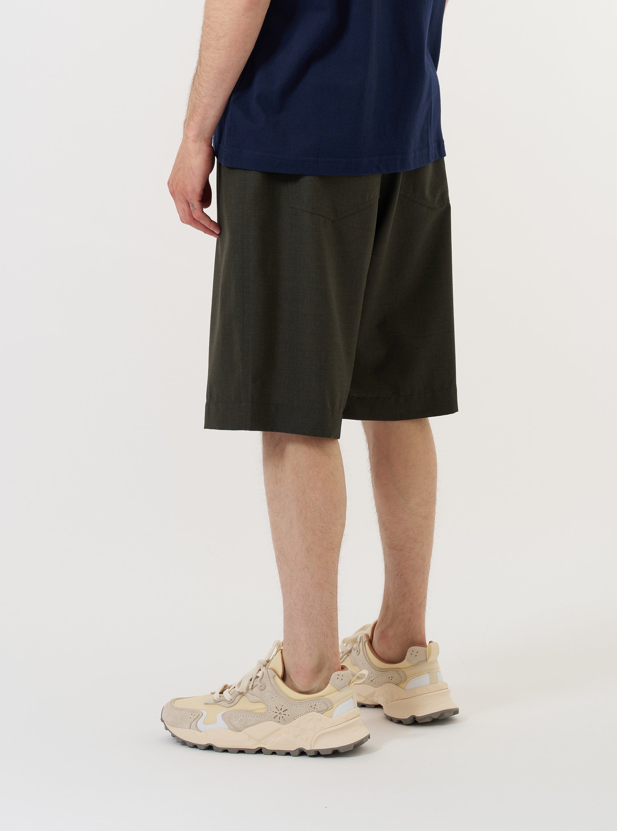A model is wearing the Olive Marl Tropical Suiting Vacation Short against a plain white background. The olive green, knee-length shorts feature a relaxed fit and a drawstring waistband, and are shown from the back to highlight the simple, clean design and the texture of the tropical suiting fabric.