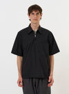 A model is wearing the Black Broadcloth Ewan Shirt against a plain white background. The shirt is a black, long-sleeved pullover featuring a pointed collar and a silver quarter-zip at the neck. The image focuses on the upper body, showcasing the crisp texture of the broadcloth fabric and the clean design of the neckline. No mannequin is present.
