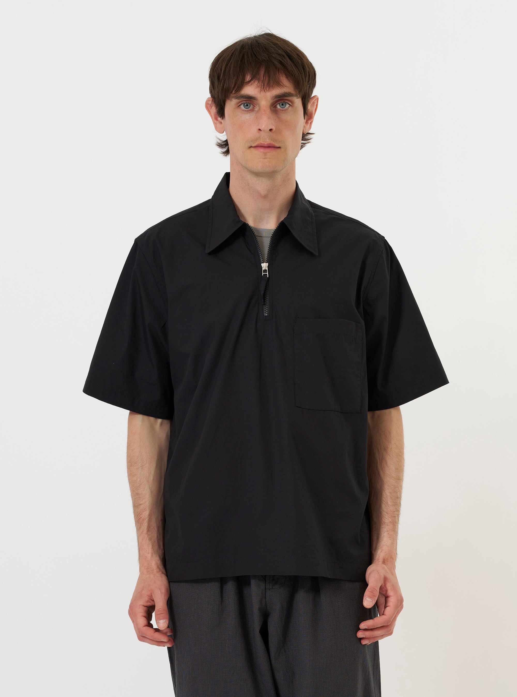 A model is wearing the Black Broadcloth Ewan Shirt against a plain white background. The shirt is a black, long-sleeved pullover featuring a pointed collar and a silver quarter-zip at the neck. The image focuses on the upper body, showcasing the crisp texture of the broadcloth fabric and the clean design of the neckline. No mannequin is present.