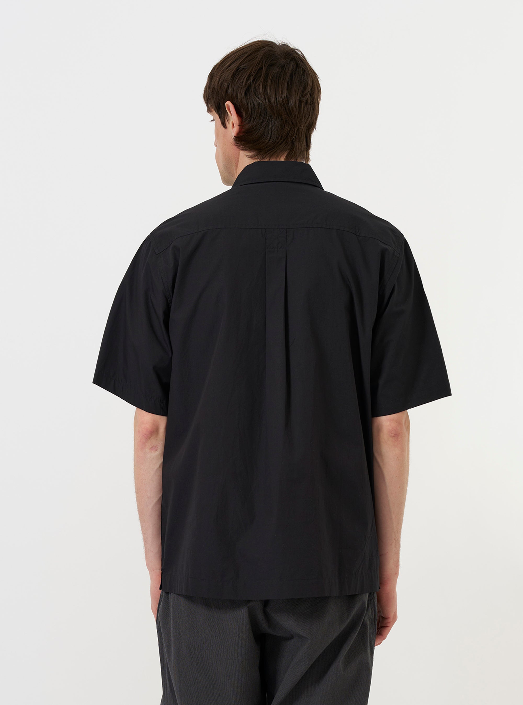 A model is shown from the back wearing the Black Broadcloth Ewan Shirt against a plain white background. The image highlights the shirt's pullover construction, classic collar, and straight hem. The model is pairing the black broadcloth garment with dark trousers.