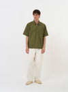The Olive Broadcloth Ewan Shirt is displayed laid flat against a solid white background. The shirt features a relaxed, wide fit with short sleeves, a classic collar, and a visible silver quarter-zip at the neck. A single patch pocket is located on the left chest, and the hem is straight with small side splits.
