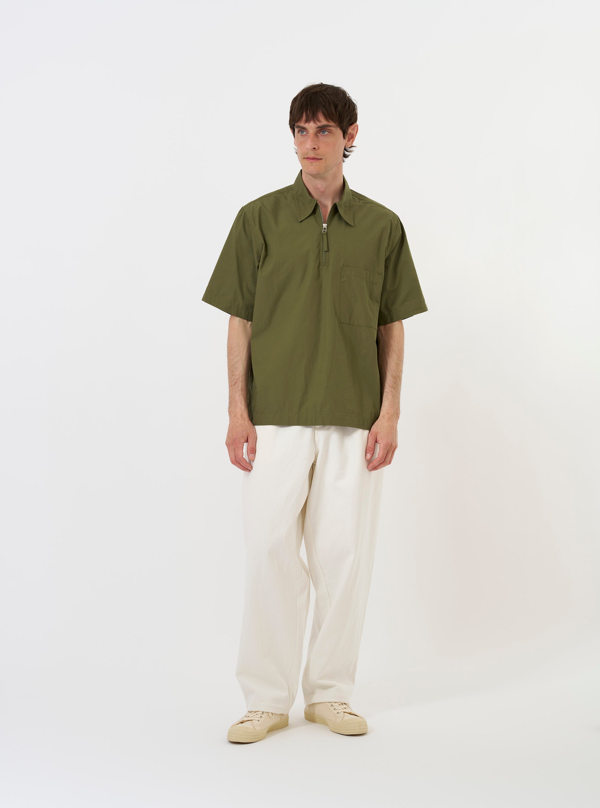 The Olive Broadcloth Ewan Shirt is displayed laid flat against a solid white background. The shirt features a relaxed, wide fit with short sleeves, a classic collar, and a visible silver quarter-zip at the neck. A single patch pocket is located on the left chest, and the hem is straight with small side splits.