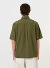 A model is wearing the Olive Broadcloth Ewan Shirt against a plain white background. The image provides a rear view of the model from the waist up, highlighting the olive green fabric, the back yoke, and the straight hem of the garment. The model is also wearing dark trousers. No mannequin is present in the image.
