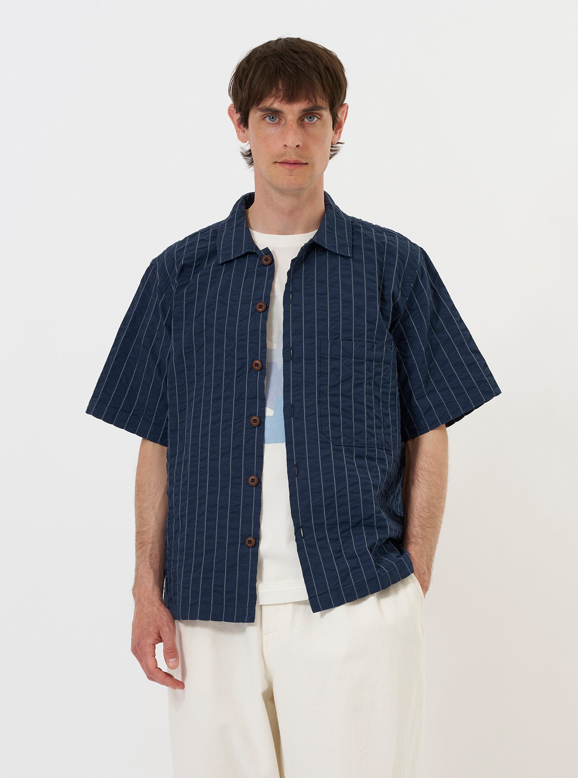 A model wears the Navy Chester Stripe S/S Fatigue Shirt against a plain white background. The short-sleeved overshirt is shown from the chest up, featuring a classic pointed collar, a button-down front, and a prominent lowered patch pocket on the left side. The fabric is a navy blue cotton seersucker with white pinstripes. The model is standing and wearing the garment as a top layer.