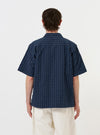 A model is wearing the Navy Chester Stripe S/S Fatigue Shirt against a plain white background. The image shows the back of the short-sleeved overshirt, highlighting the navy blue and white pinstriped seersucker fabric, a straight hem, and a simple collar. The model is standing and pairing the shirt with olive green trousers. No mannequin is present.