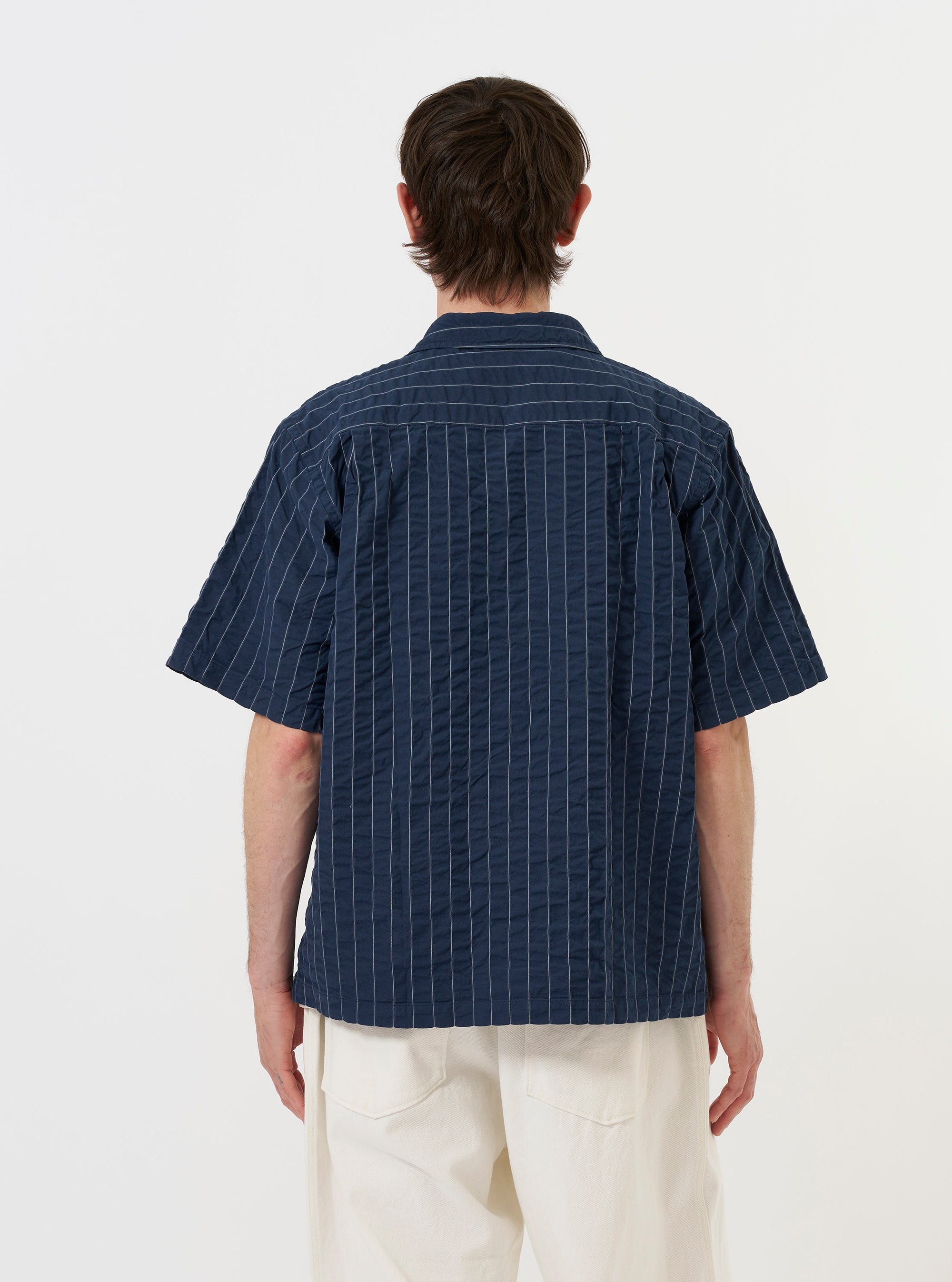 A model is wearing the Navy Chester Stripe S/S Fatigue Shirt against a plain white background. The image shows the back of the short-sleeved overshirt, highlighting the navy blue and white pinstriped seersucker fabric, a straight hem, and a simple collar. The model is standing and pairing the shirt with olive green trousers. No mannequin is present.