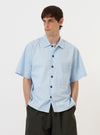 A model is wearing the Sky Blue Paper Touch Cotton S/S Fatigue Shirt, standing against a plain white background. The shirt features a loose fit, short sleeves, a simple pointed collar, and a single patch pocket positioned lower on the left chest. The sky blue cotton fabric appears crisp and structured, and the model is shown from the waist up, highlighting the garment's fit and texture.