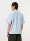 A model is wearing the Sky Blue Paper Touch Cotton S/S Fatigue Shirt against a plain white background. The image provides a close-up rear view of the garment, highlighting the straight hem, short sleeves, and the crisp texture of the sky blue fabric. The model is also wearing dark-colored trousers.