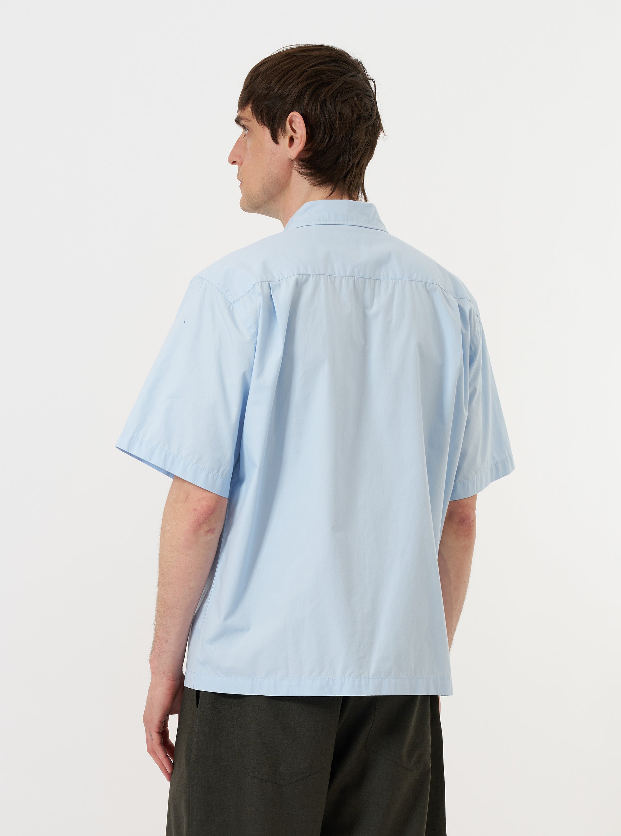 A model is wearing the Sky Blue Paper Touch Cotton S/S Fatigue Shirt against a plain white background. The image provides a close-up rear view of the garment, highlighting the straight hem, short sleeves, and the crisp texture of the sky blue fabric. The model is also wearing dark-colored trousers.