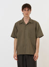 A model is wearing the Olive Linen Cotton Shirting Camp Shirt against a plain white background. The shirt features an olive green color, a relaxed camp collar, a button-down front, and short sleeves. The model is shown from the chest up, with the shirt worn open over a white t-shirt, highlighting the texture of the linen-cotton fabric.