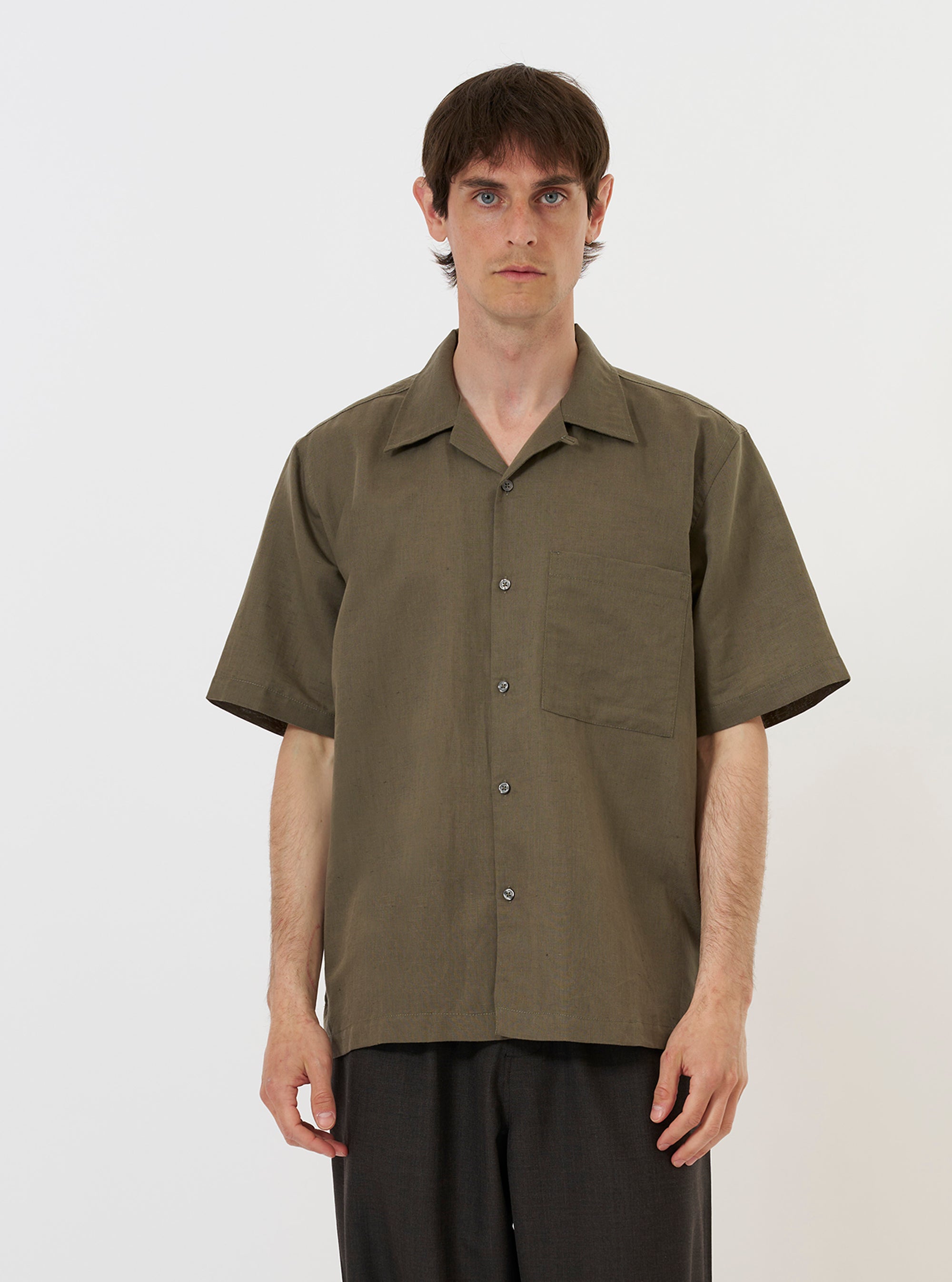 A model is wearing the Olive Linen Cotton Shirting Camp Shirt against a plain white background. The shirt features an olive green color, a relaxed camp collar, a button-down front, and short sleeves. The model is shown from the chest up, with the shirt worn open over a white t-shirt, highlighting the texture of the linen-cotton fabric.