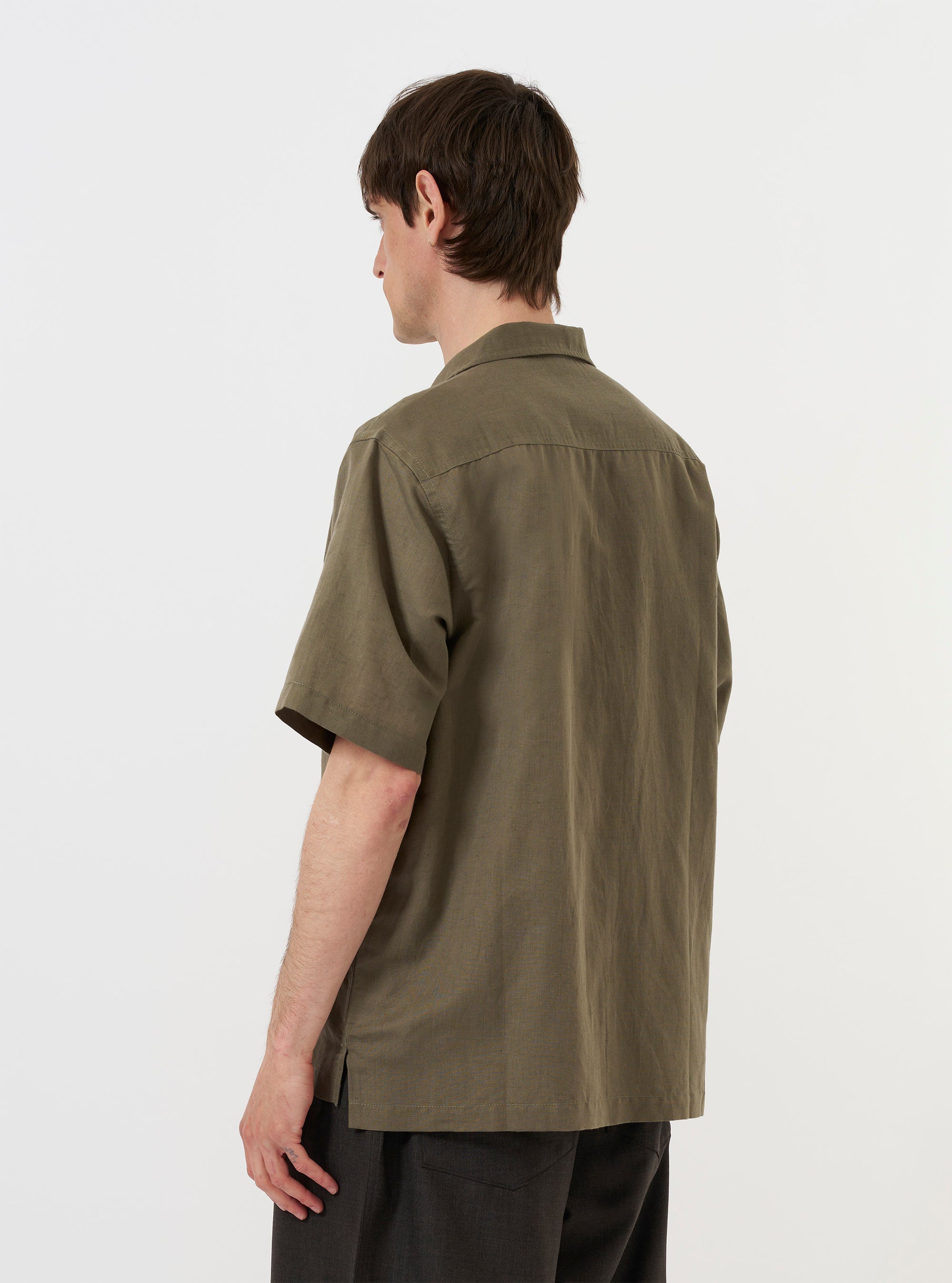 A model is wearing the Olive Linen Cotton Shirting Camp Shirt against a plain white background. The image shows the shirt from the back, highlighting the yoke with small pleats and the elongated hem with side splits. The model is standing with their hands in their pockets, and the olive green shirt is paired with light-colored trousers.