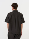 A male model is shown from the back wearing the Brown Marl Tropical Suiting Ewan Shirt against a plain white background. The image highlights the short sleeves, the straight hem with side splits, the back yoke, and the central box pleat. The model is also wearing olive green trousers.