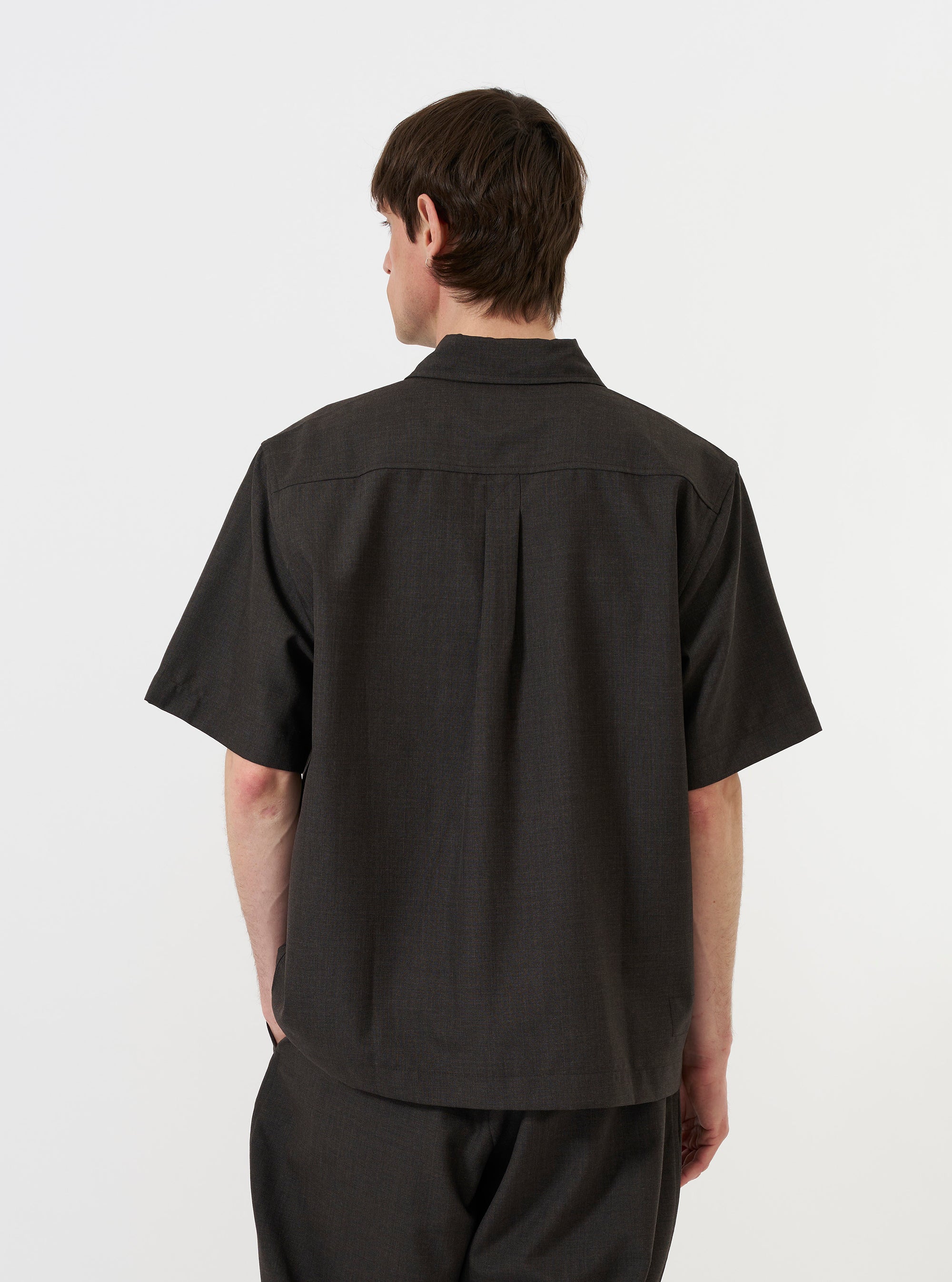 A male model is shown from the back wearing the Brown Marl Tropical Suiting Ewan Shirt against a plain white background. The image highlights the short sleeves, the straight hem with side splits, the back yoke, and the central box pleat. The model is also wearing olive green trousers.