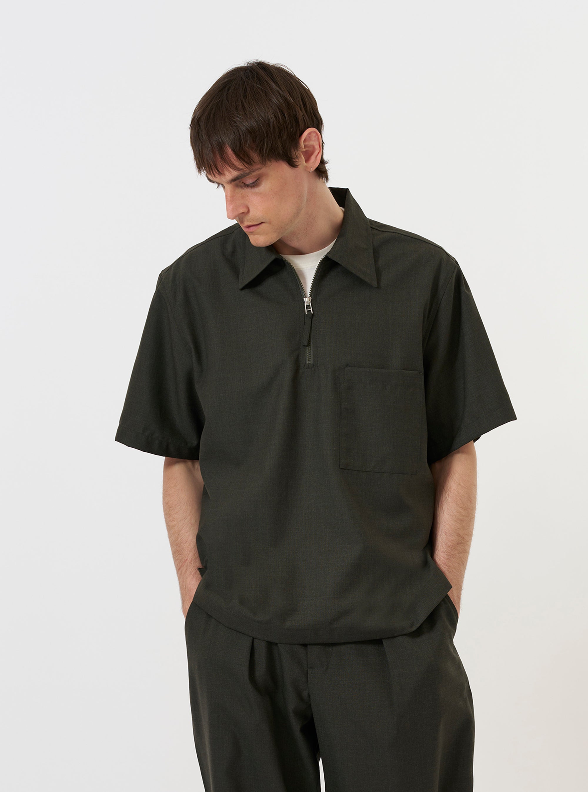 A model is wearing the Olive Marl Tropical Suiting Ewan Shirt against a plain white background. The short-sleeved shirt is shown from the front with the quarter-zip neckline partially open, highlighting its relaxed fit and olive green marl fabric.